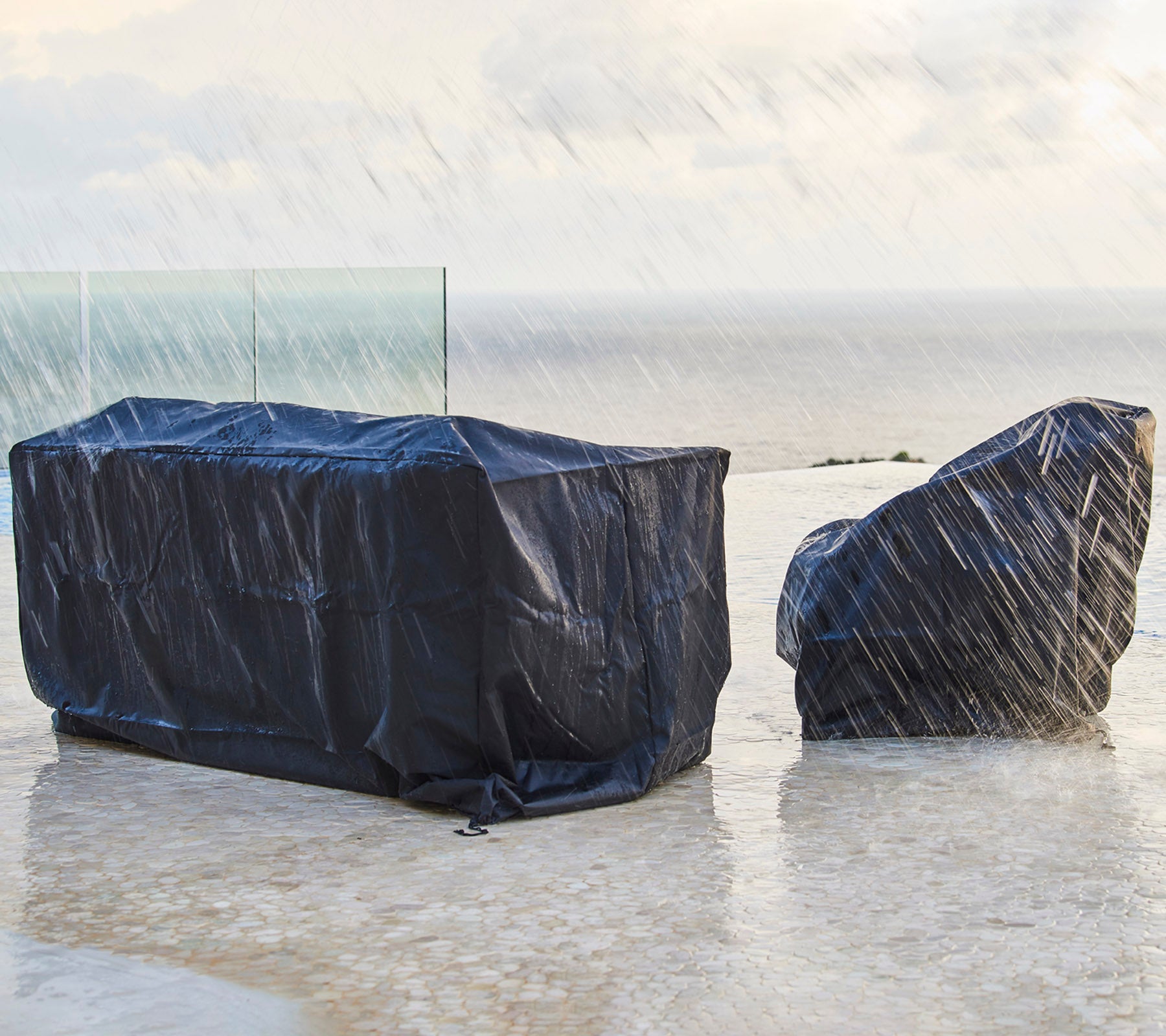 Muebles de exterior cubiertos con fundas protectoras oscuras, a salvo de la lluvia, con un fondo de un cielo nublado y el océano.