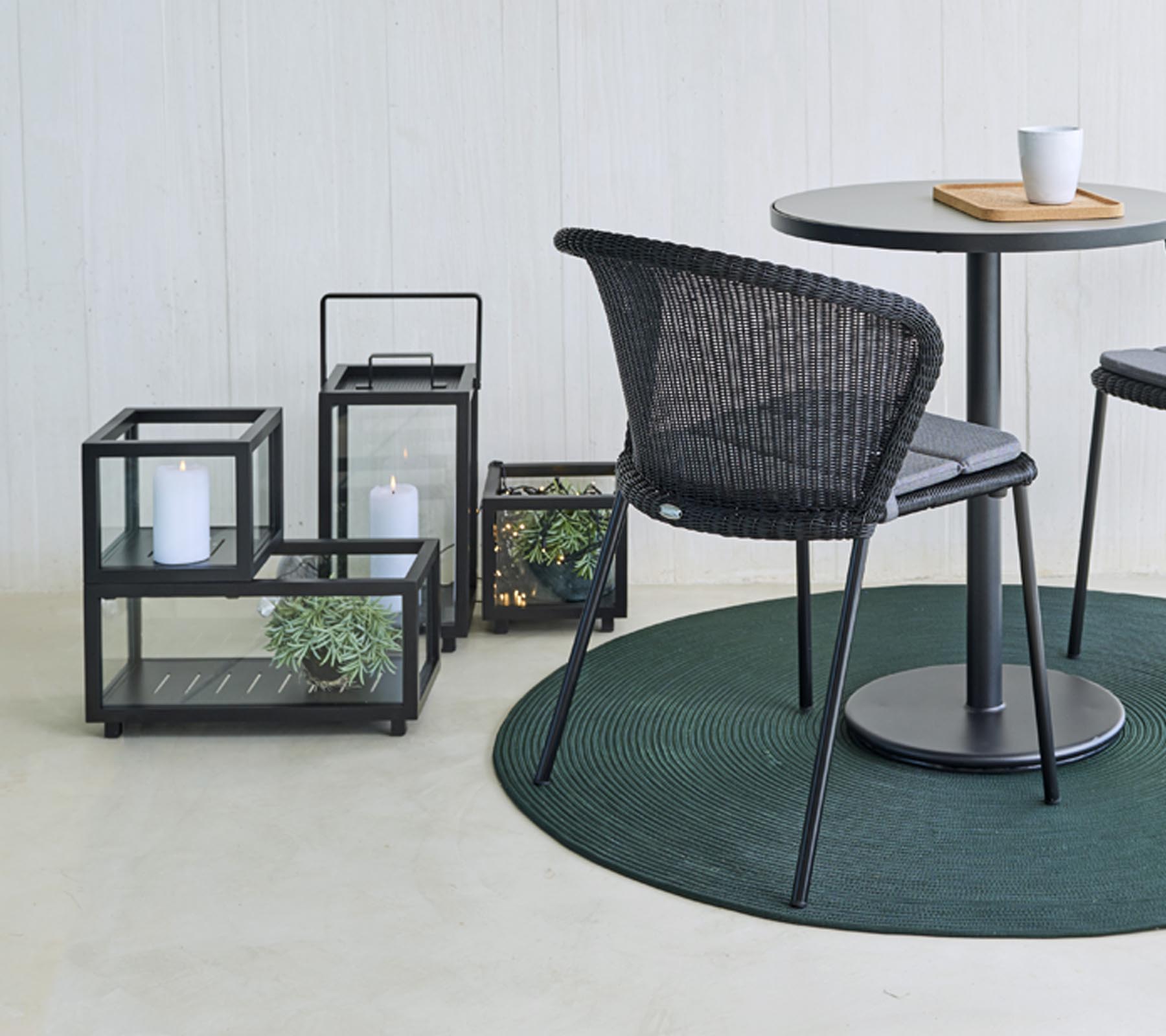 Asientos modernos y faroles de vidrio decorativos con plantas, dispuestos alrededor de una mesa redonda, sobre una alfombra redonda verde.