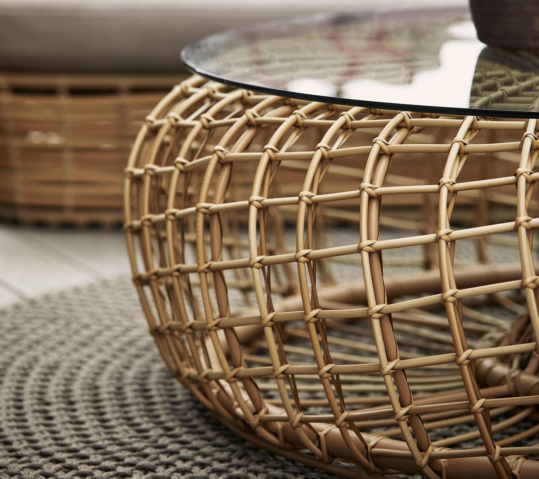 Mesa de café redonda con una base de ratán trenzado y una tapa de vidrio, complementada por una alfombra circular texturizada.