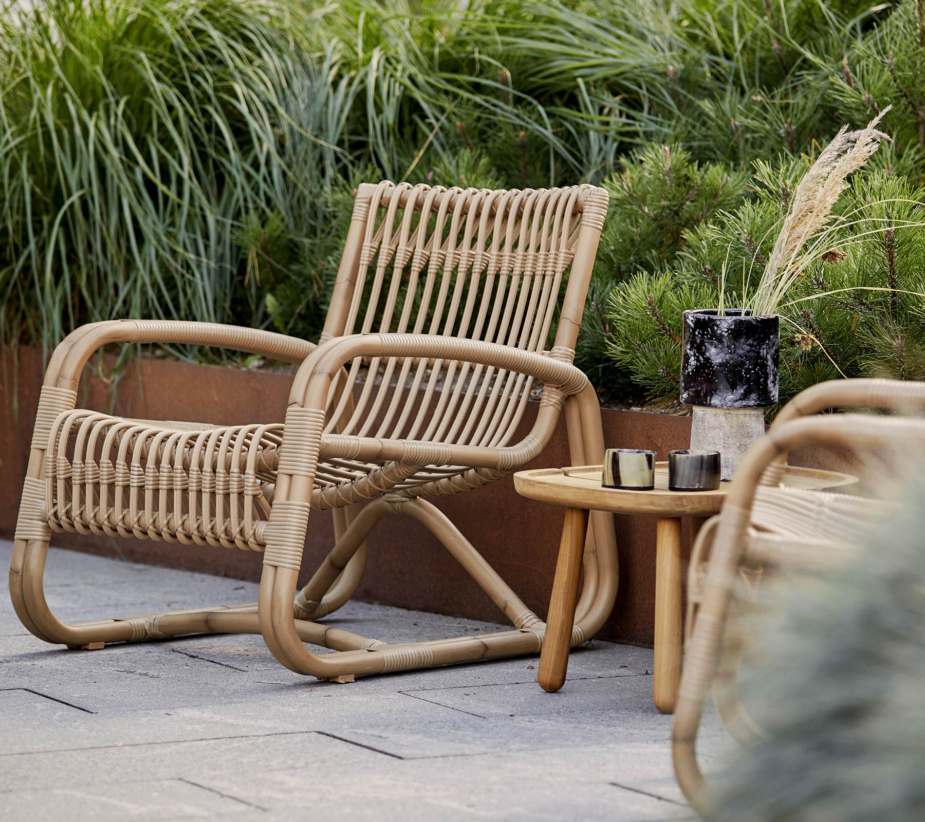 Una silla tejida de forma natural acompañada de una pequeña mesa de madera y elementos decorativos, rodeada de un exuberante verdor.