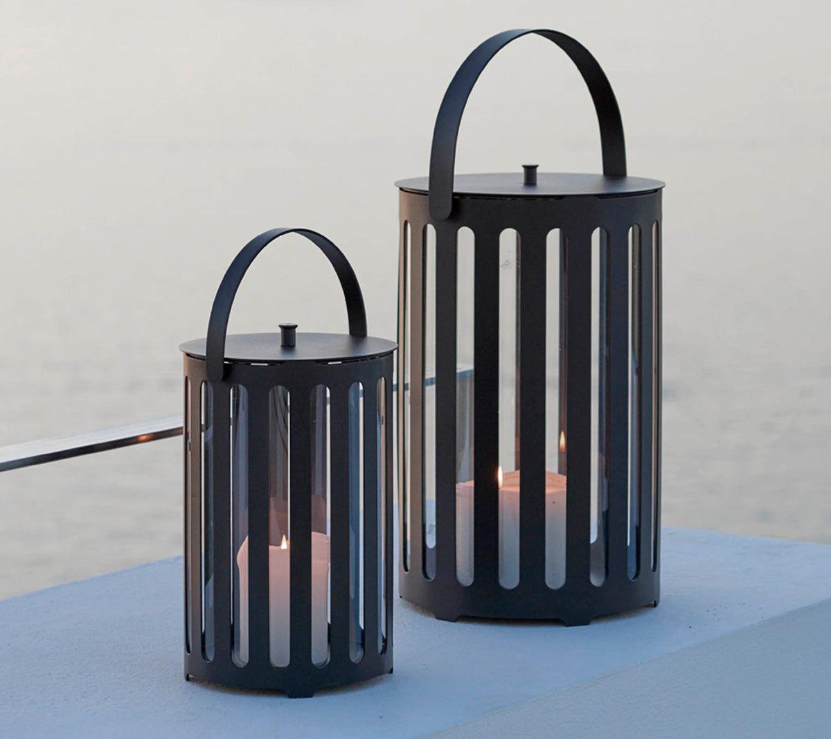 Dos elegantes faroles de metal con ranuras verticales, que cuentan con asas y velas en su interior, contra un fondo sereno.
