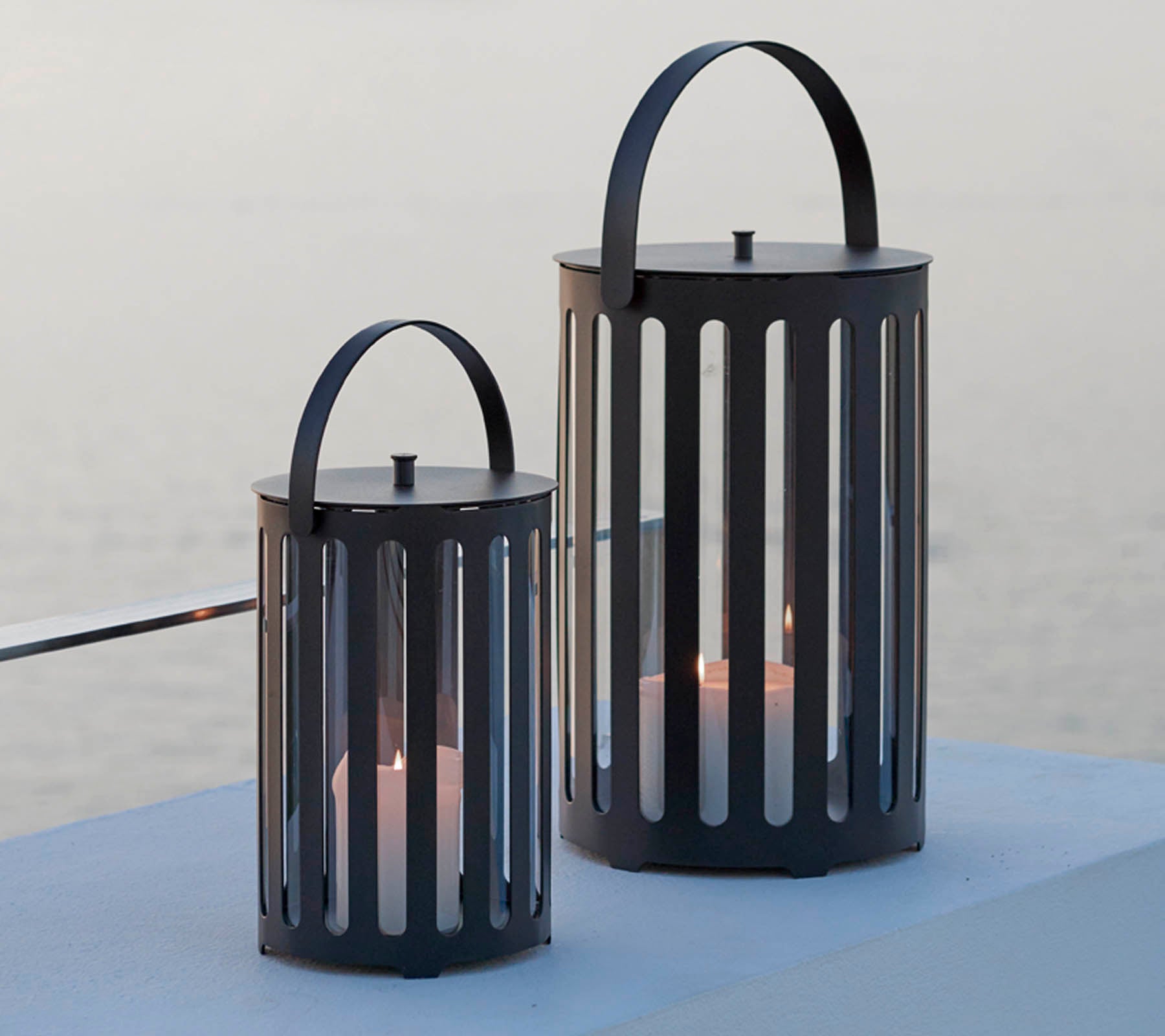 Dos elegantes faroles de metal con ranuras verticales, que cuentan con asas y velas en su interior, contra un fondo sereno.