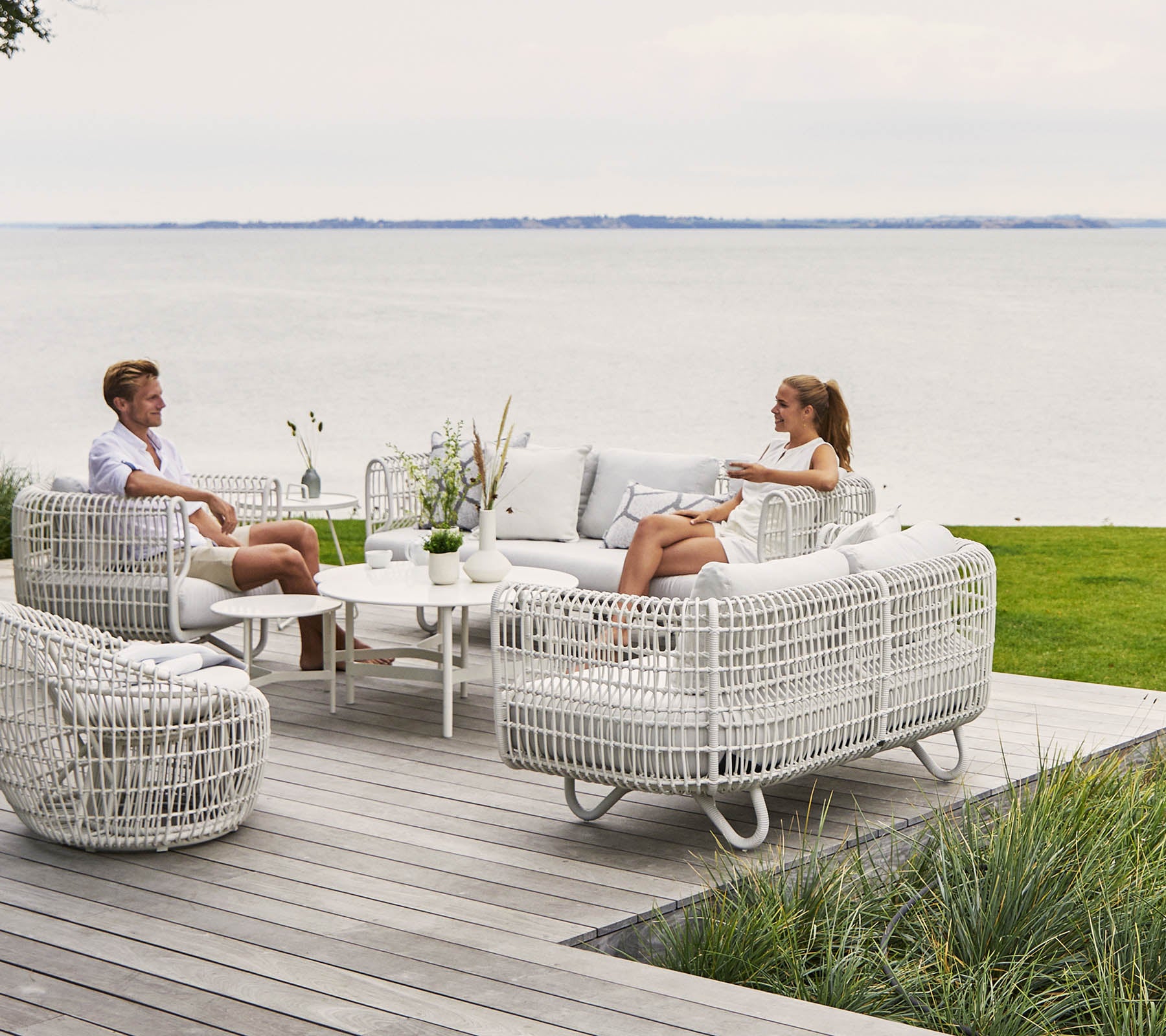 Dos personas se relajan en una elegante y cómoda área de descanso con muebles de mimbre blanco y una mesa de café, con vistas a una pintoresca vista del agua.