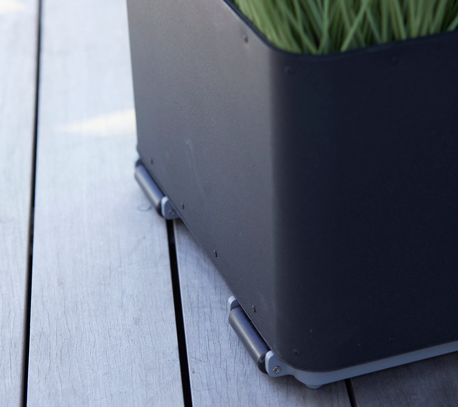 Caja de plantas negra con un diseño elegante, que presenta césped verde y conectores plateados sobre una superficie de madera.