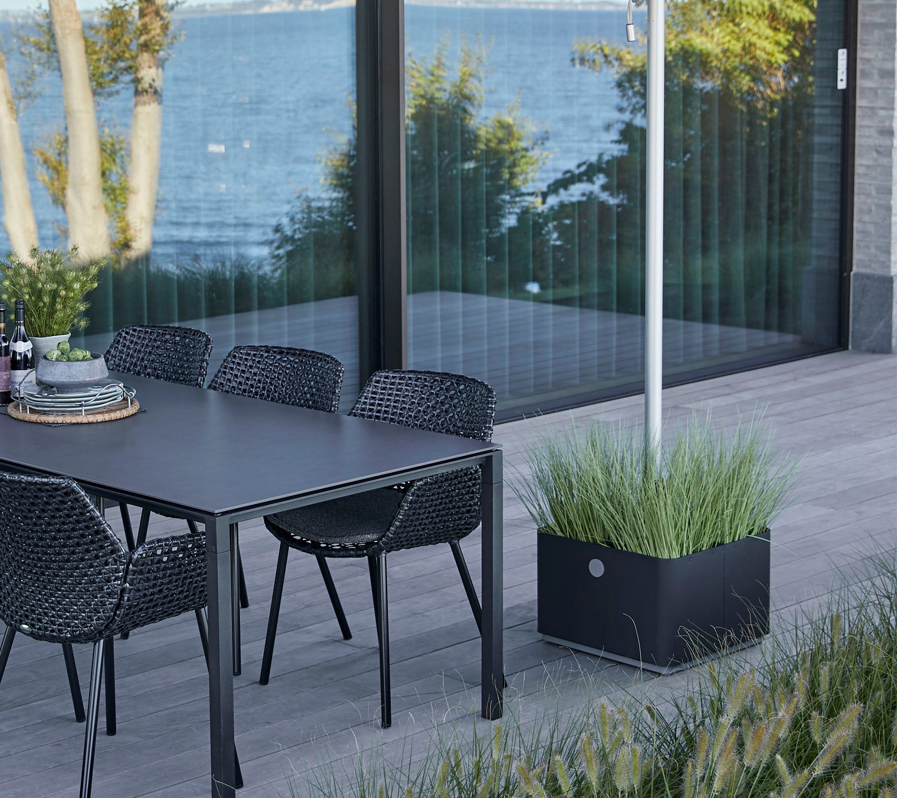 Una mesa elegante con sillas colocadas junto a una vista al agua, con un jardinero decorativo con hierba exuberante.