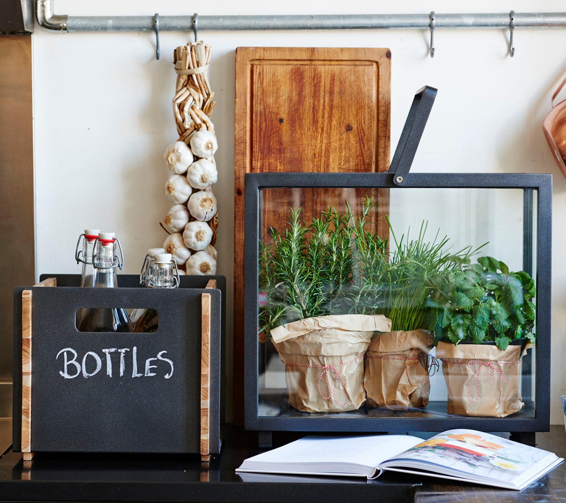 Una exhibición elegante que presenta una caja negra etiquetada 'BOTELLAS', un recipiente de vidrio con hierbas en macetas y un libro de cocina sobre una superficie.
