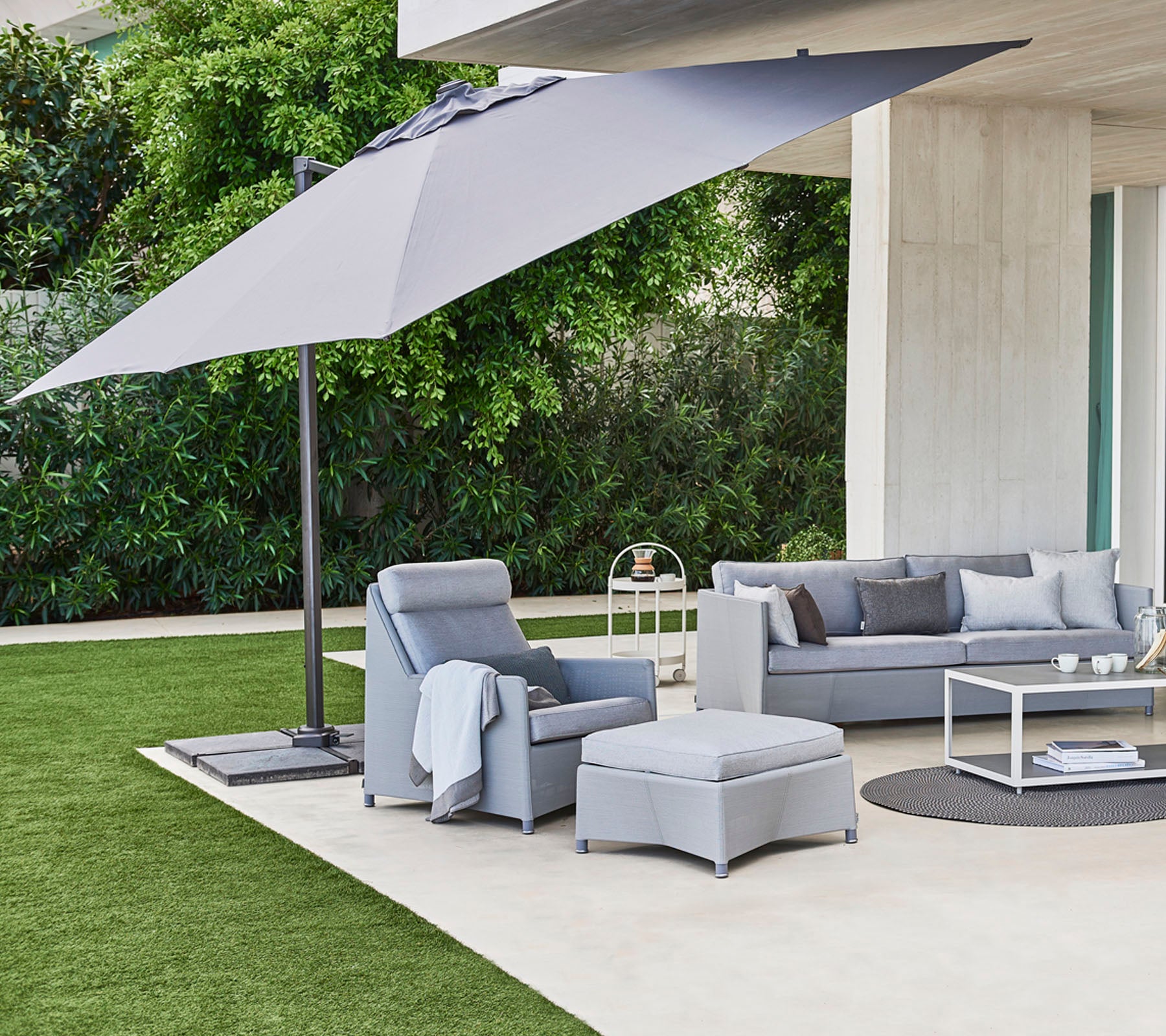 Asientos exteriores elegantes con cojines acolchados, una mesa de café y una gran sombrilla que proporciona sombra contra un fondo verde.