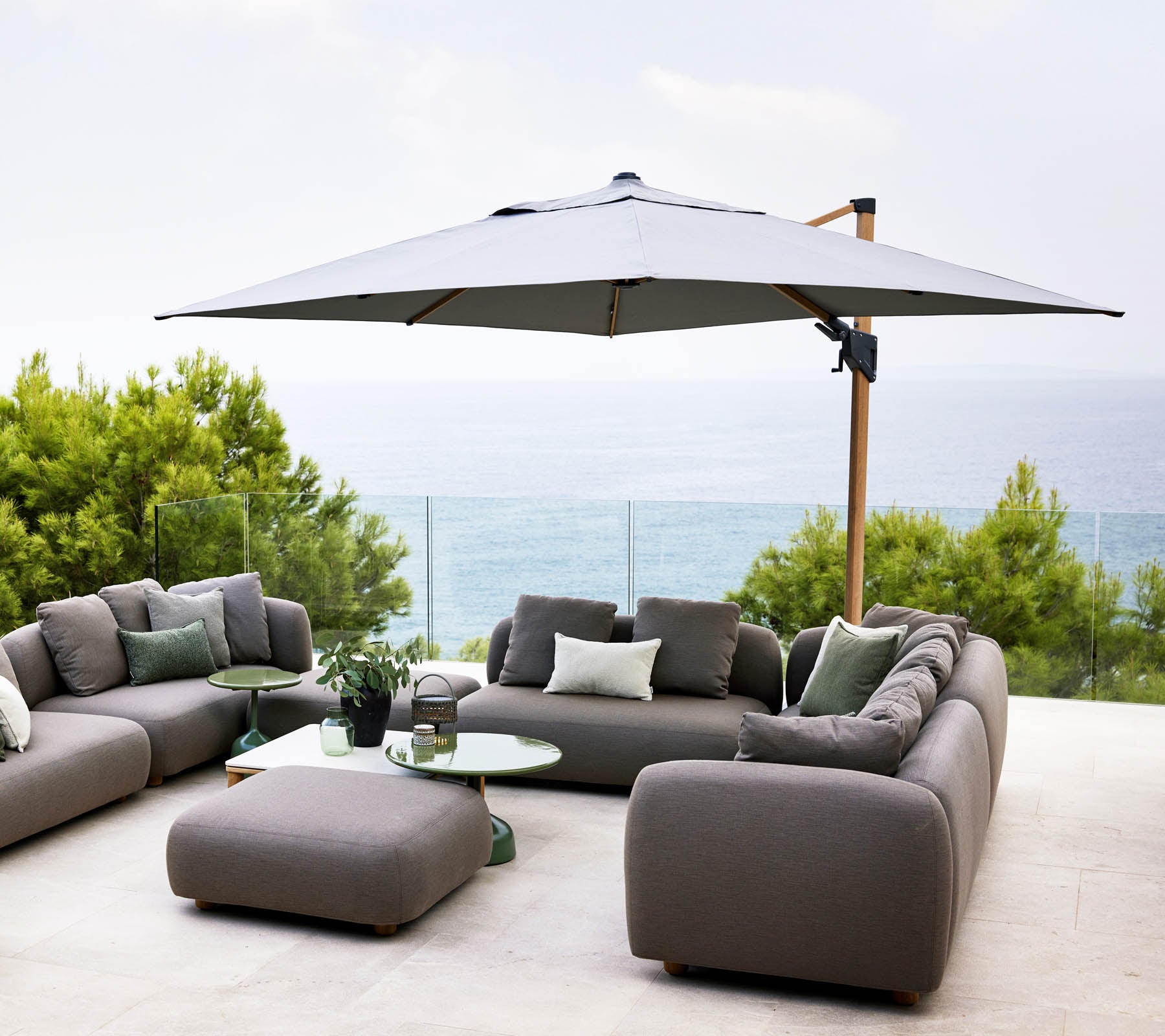 Área de estar al aire libre elegante con una gran sombrilla, sofás grises y una mesa de vidrio, con vistas a una serena vista del agua y vegetación exuberante.