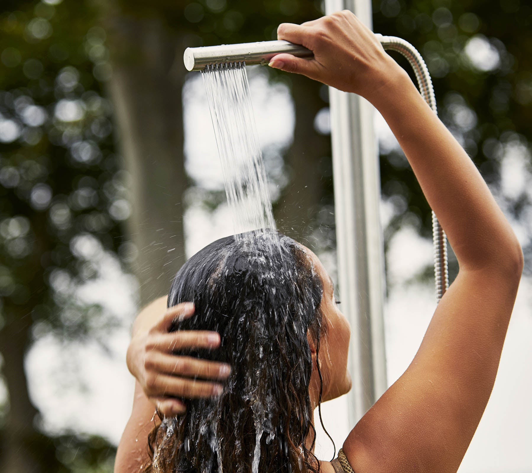 Una persona lavándose el cabello bajo la ducha, con el agua cayendo y rodeada de un entorno natural.