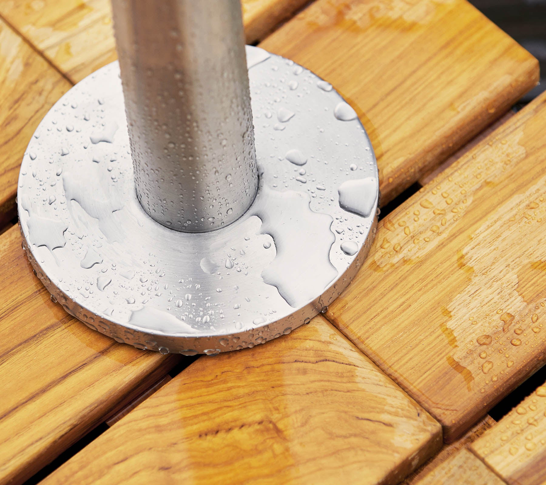 Base de metal con gotas de agua descansando sobre una superficie de madera, con tablones lisos y ranurados y un poste cilíndrico en el centro.