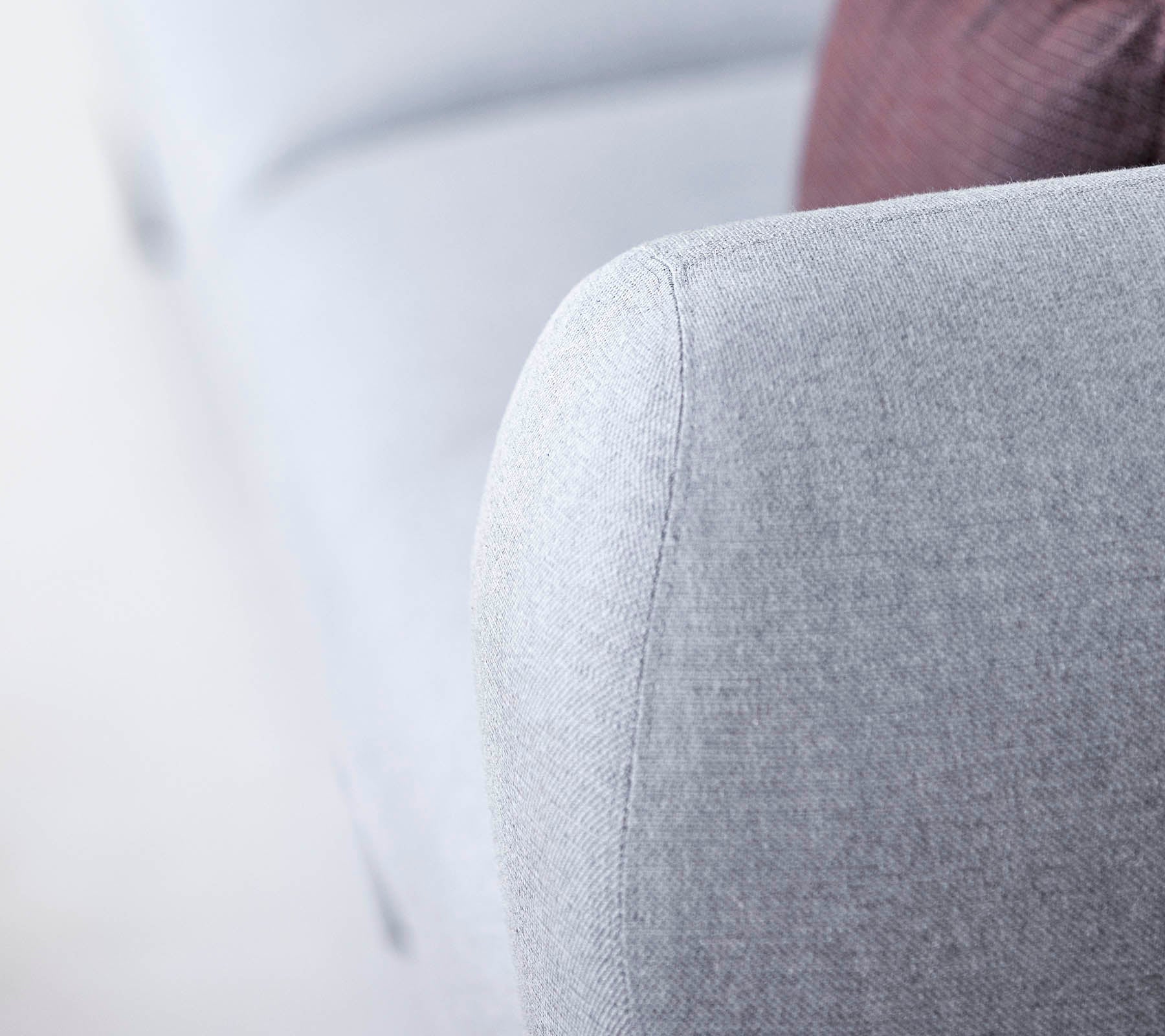 Acercamiento de un brazo de sofá gris claro, con una tela texturizada y un diseño suave y redondeado, con un toque de una almohada más oscura en el fondo.
