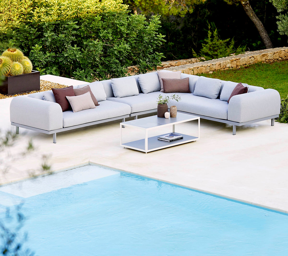 Sofá seccional gris claro con cojines decorativos y una mesa de café de vidrio, situado junto a una piscina y una vegetación exuberante.
