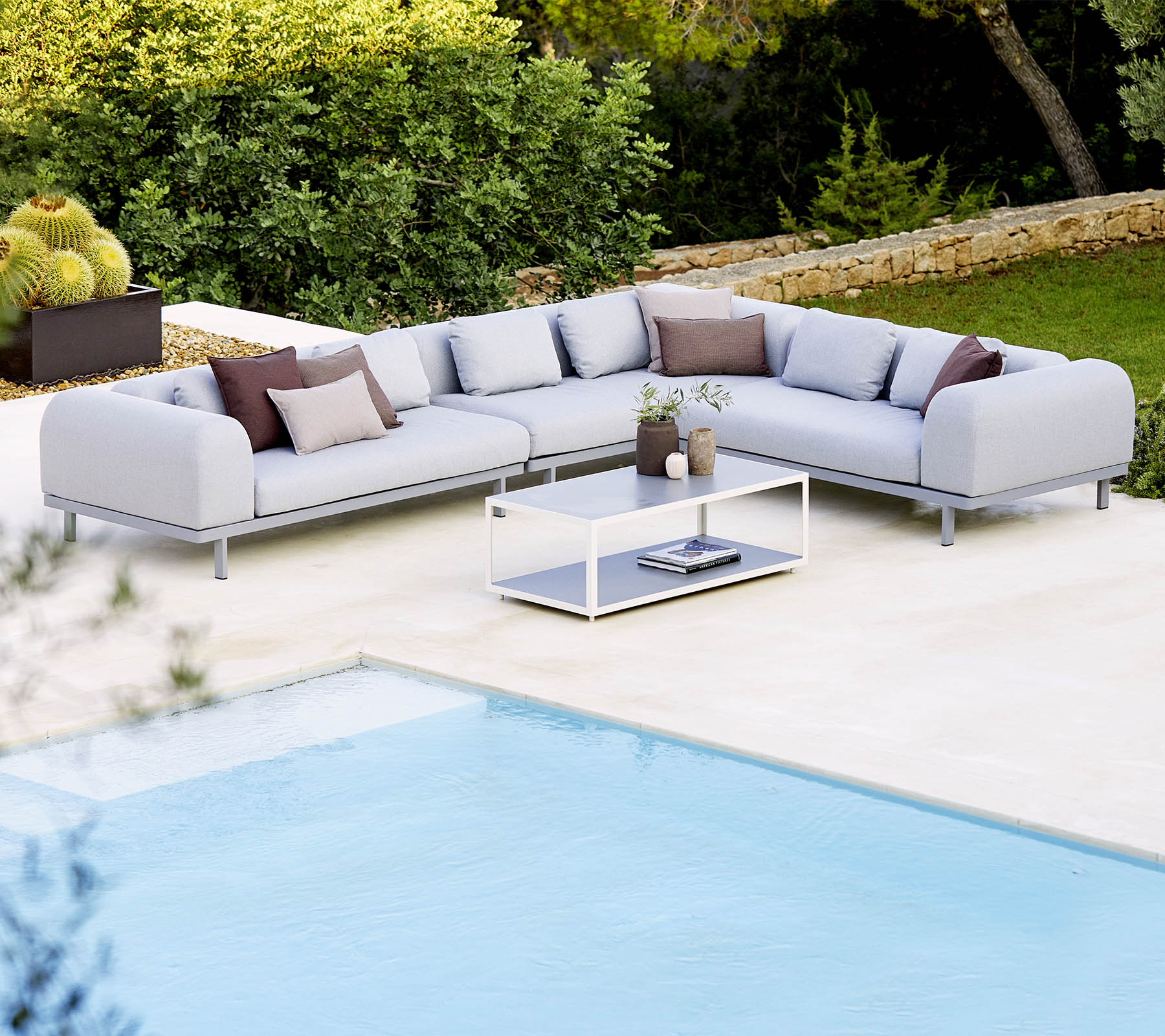 Sofá seccional gris claro con cojines decorativos y una mesa de café de vidrio, situado junto a una piscina y una vegetación exuberante.