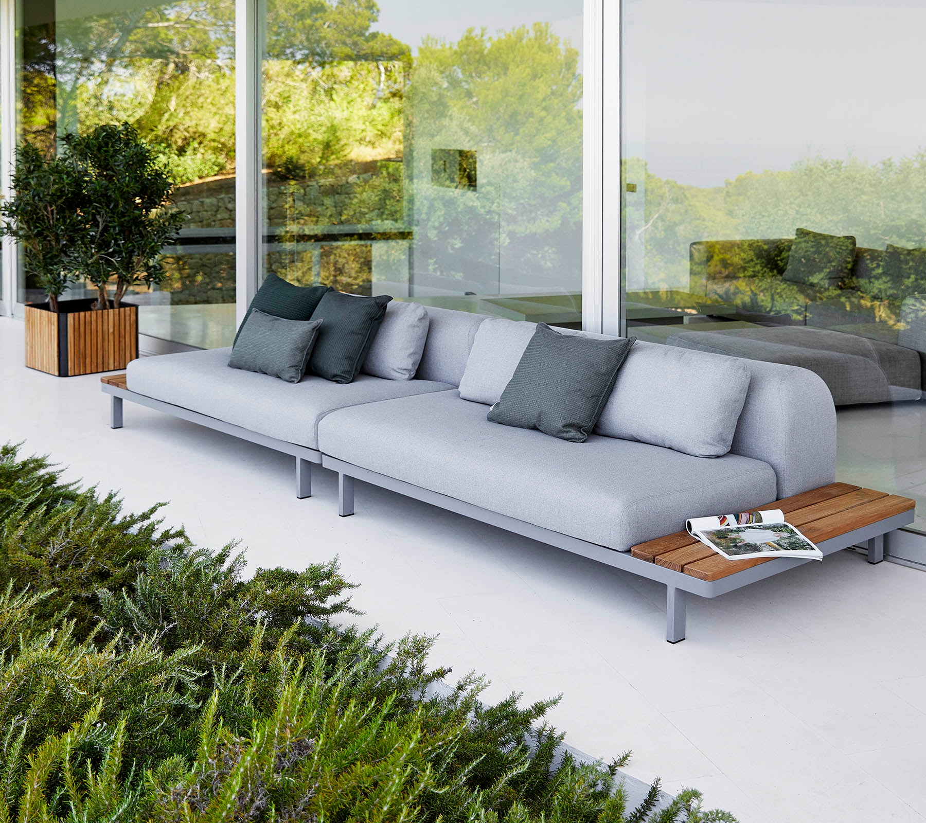Sofá moderno con cojines gris claro y acentos de madera, acompañado de cojines decorativos y rodeado de vegetación.