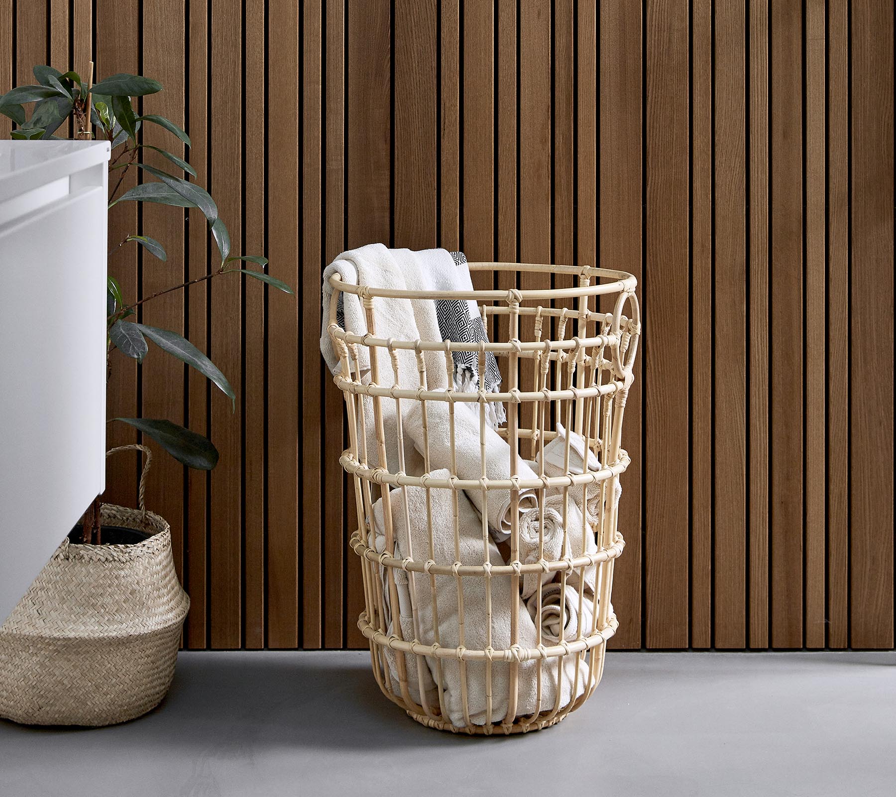 Cesta de lavandería de mimbre llena de toallas enrolladas, colocada junto a una planta y una pared de madera texturizada.