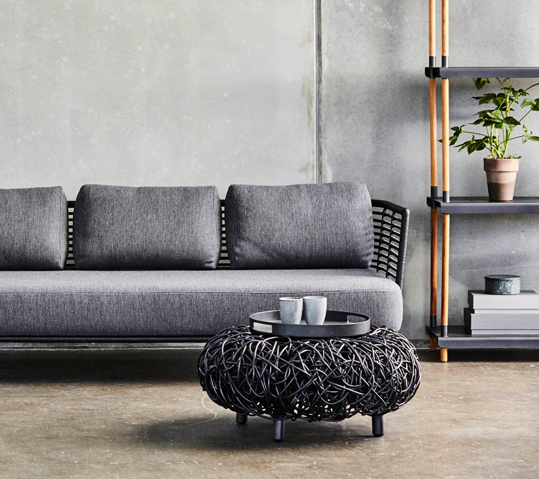 Un sofá gris moderno con cojines, combinado con una mesa de café circular de mimbre negro y una estantería minimalista que exhibe plantas y decoraciones.