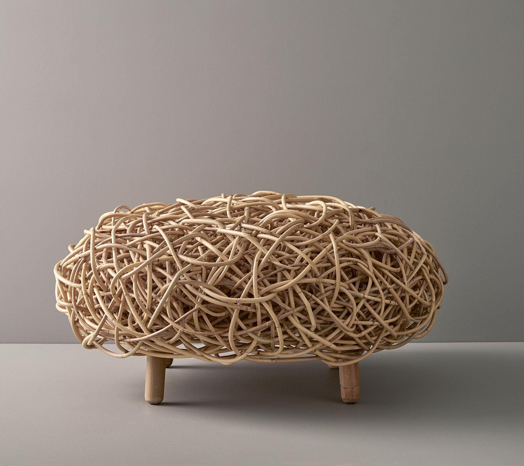 Pieza decorativa tejida de forma natural, con una forma ovalada, con hilos entrelazados intrincados y sostenida por patas de madera cortas.