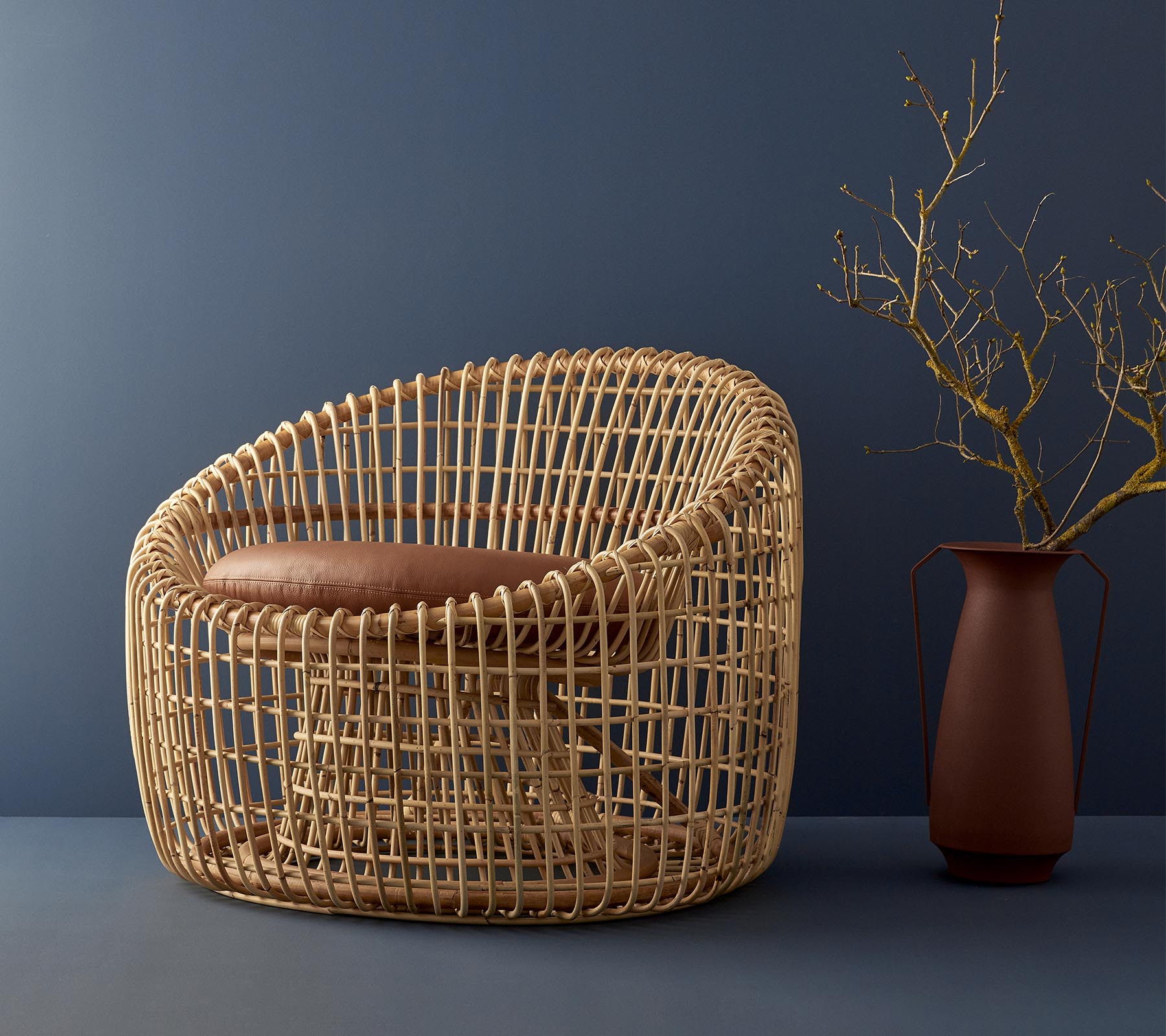 Silla moderna de mimbre con un diseño redondeado, con un asiento acolchado y complementada por un jarrón simple con una ramita.