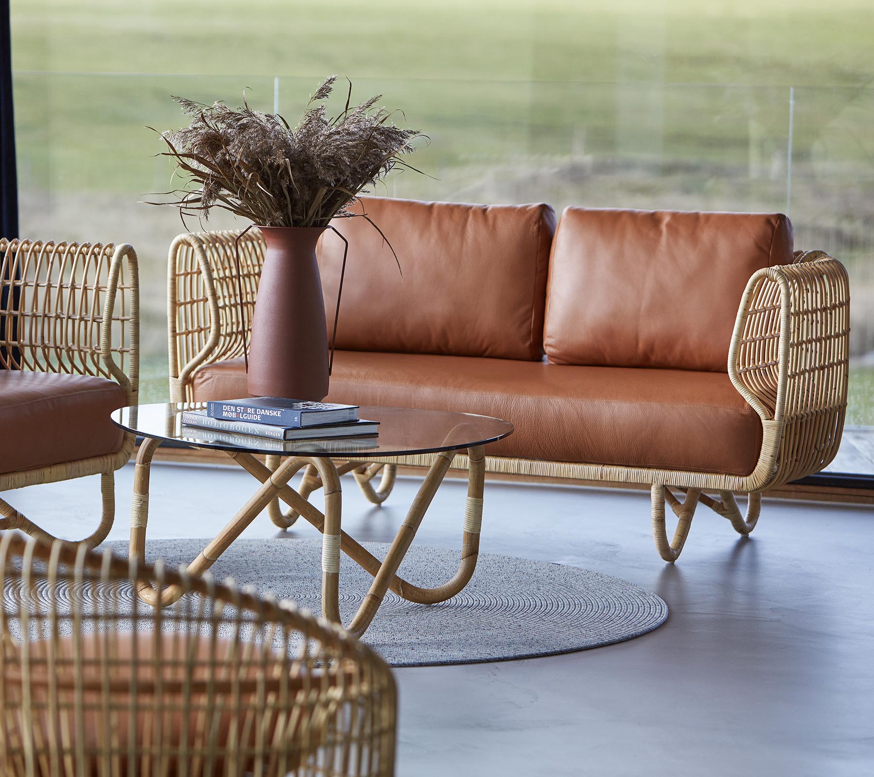 Un área de estar elegante con un sofá de cuero marrón, una mesa de café redonda y elementos decorativos como un jarrón con flores secas.