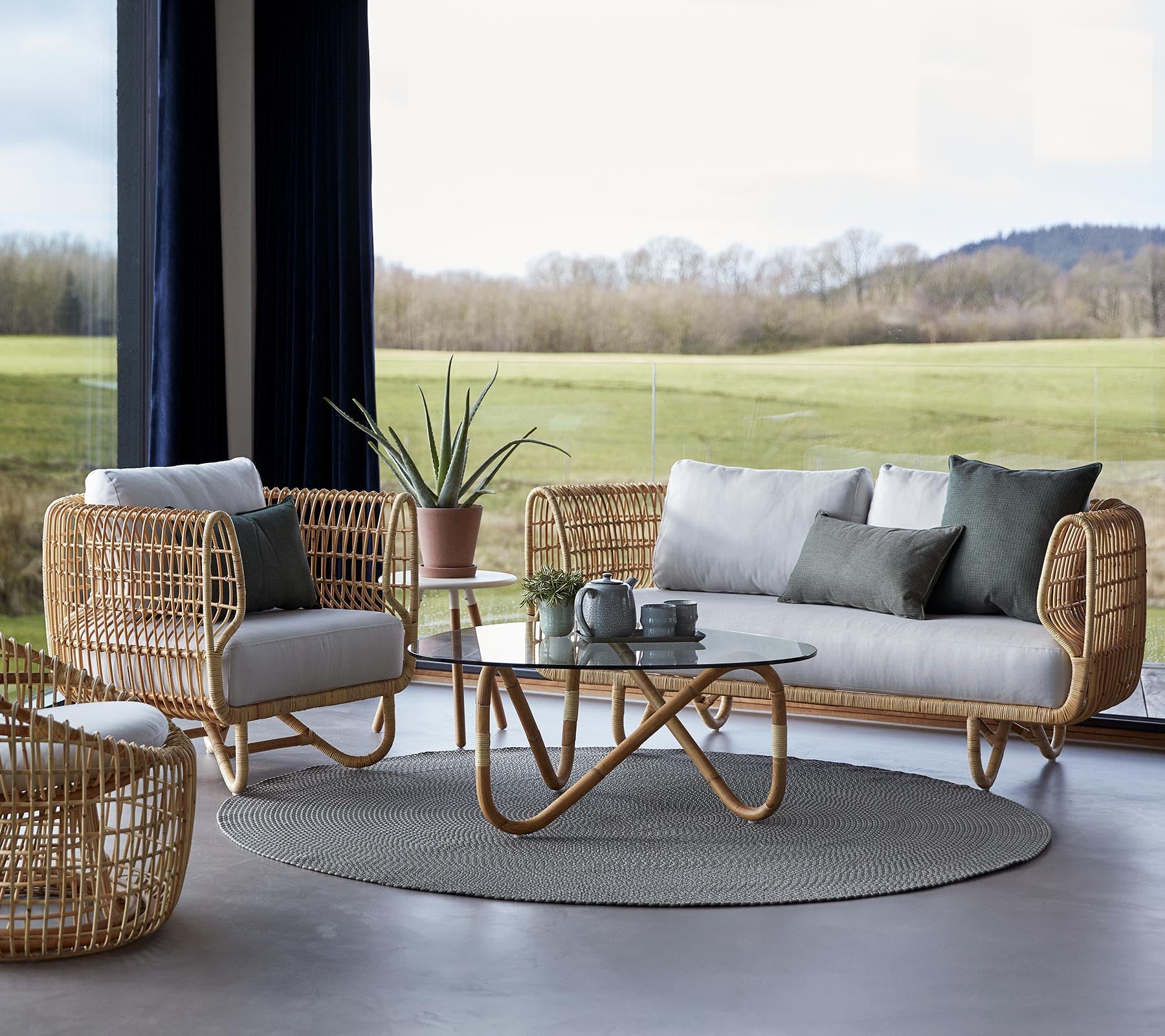 Un arreglo de asientos elegante con muebles tejidas, una alfombra redonda y una mesa de café de vidrio, complementado por elementos de decoración minimalistas.