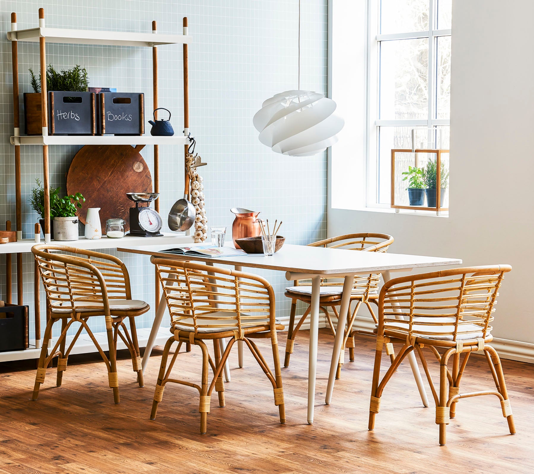 Un elegante comedor con una mesa de madera clara rodeada de sillas de ratán y una lámpara colgante moderna, complementado con estanterías decorativas.