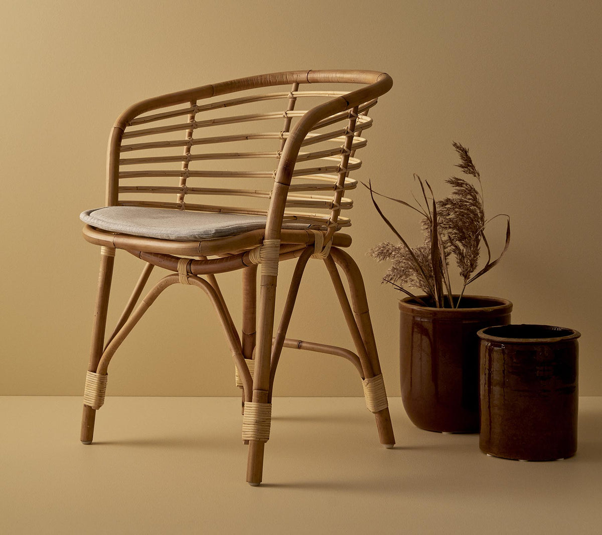 Una silla de ratán elegante con un asiento acolchado, acompañada de dos macetas decorativas con plantas secas.