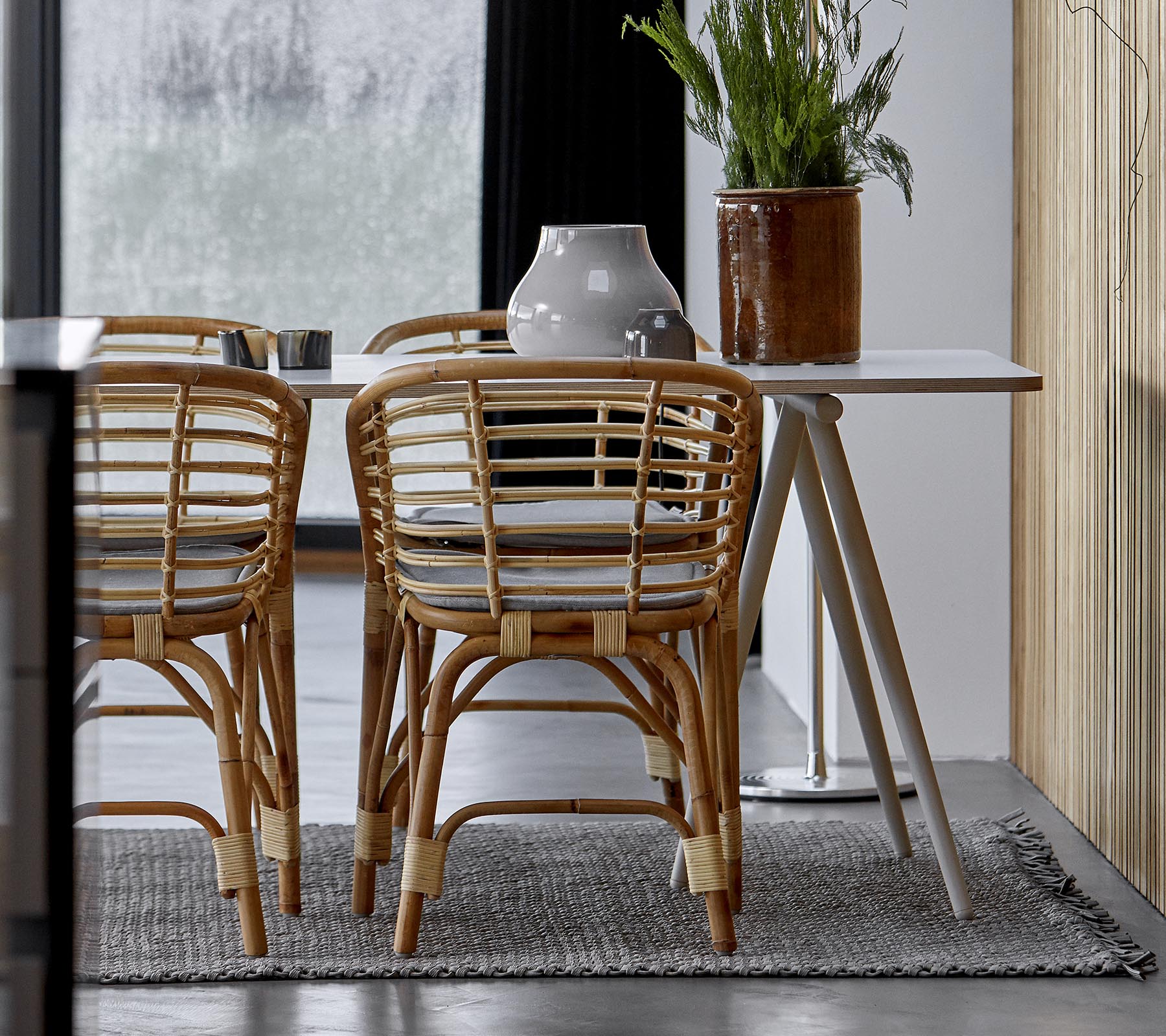 Una disposición moderna de comedor con sillas de ratán, una mesa elegante y vegetación decorativa en un interior con estilo.