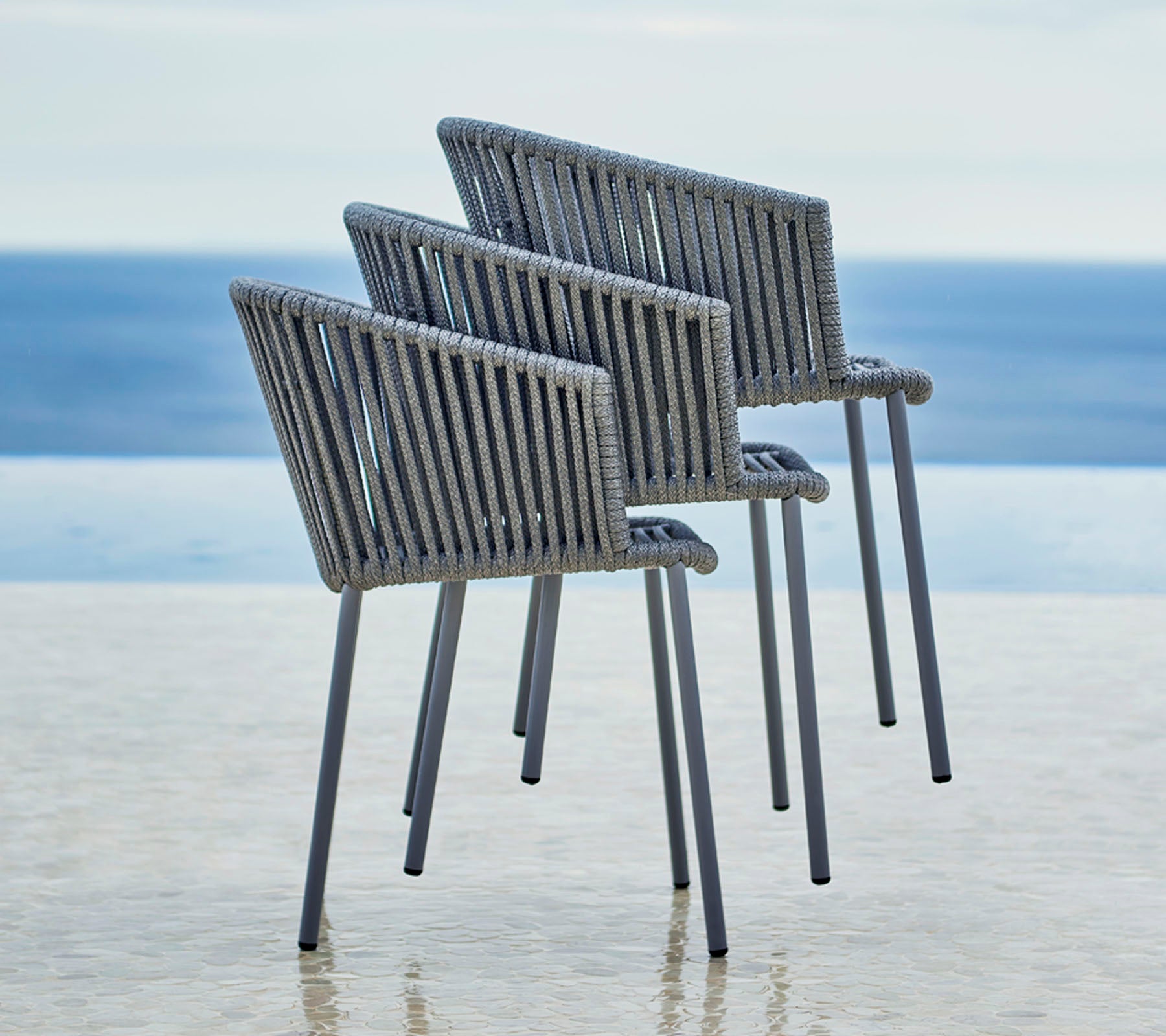 Tres sillas modernas con un diseño elegante, que cuentan con un respaldo de listones y reposabrazos, situadas junto a una serena vista al agua.
