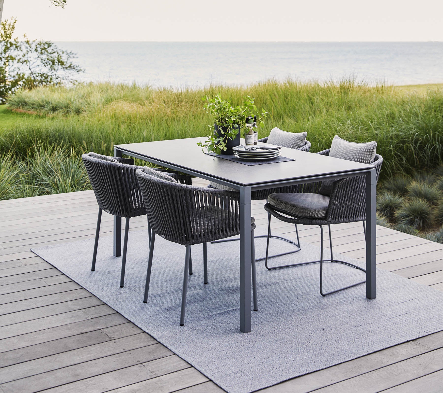 Un conjunto de comedor al aire libre moderno que cuenta con una mesa rectangular y cuatro sillas elegantes, rodeado de vegetación y una vista tranquila del agua.