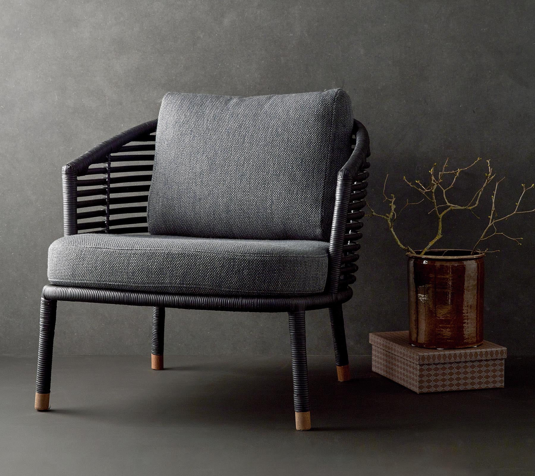 Silla elegante con un cojín gris texturizado y un marco negro trenzado, complementada con una rama decorativa y una mesa pequeña.