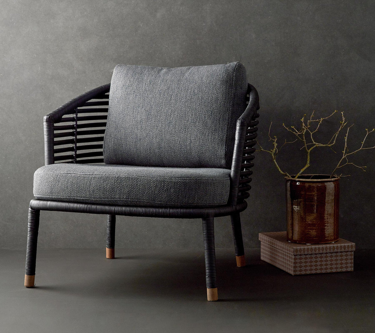 Silla elegante con un cojín gris texturizado y un marco negro trenzado, complementada con una rama decorativa y una mesa pequeña.
