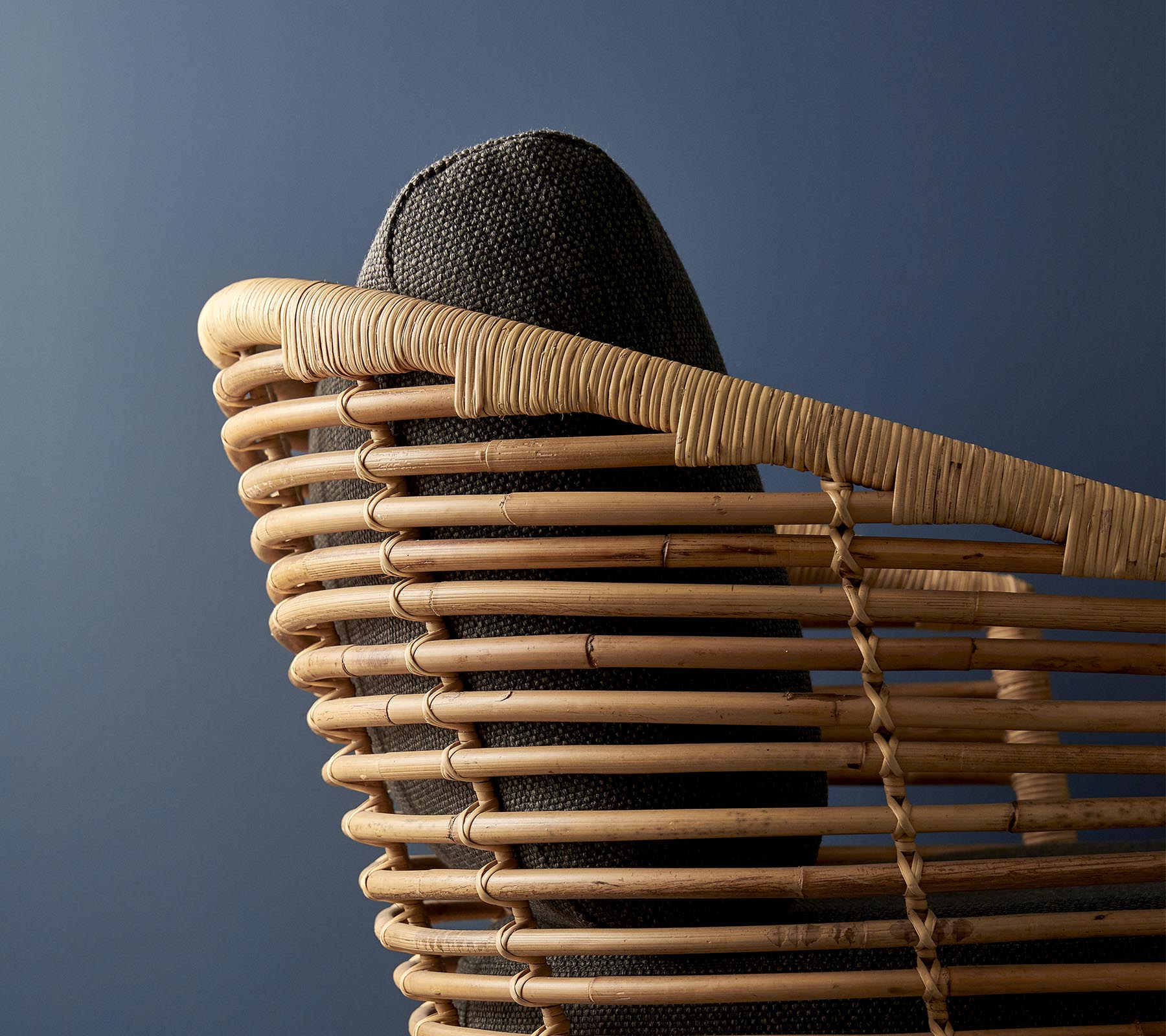 Silla tejida elegante con un respaldo curvado y un asiento acolchado oscuro contra un fondo azul apagado.