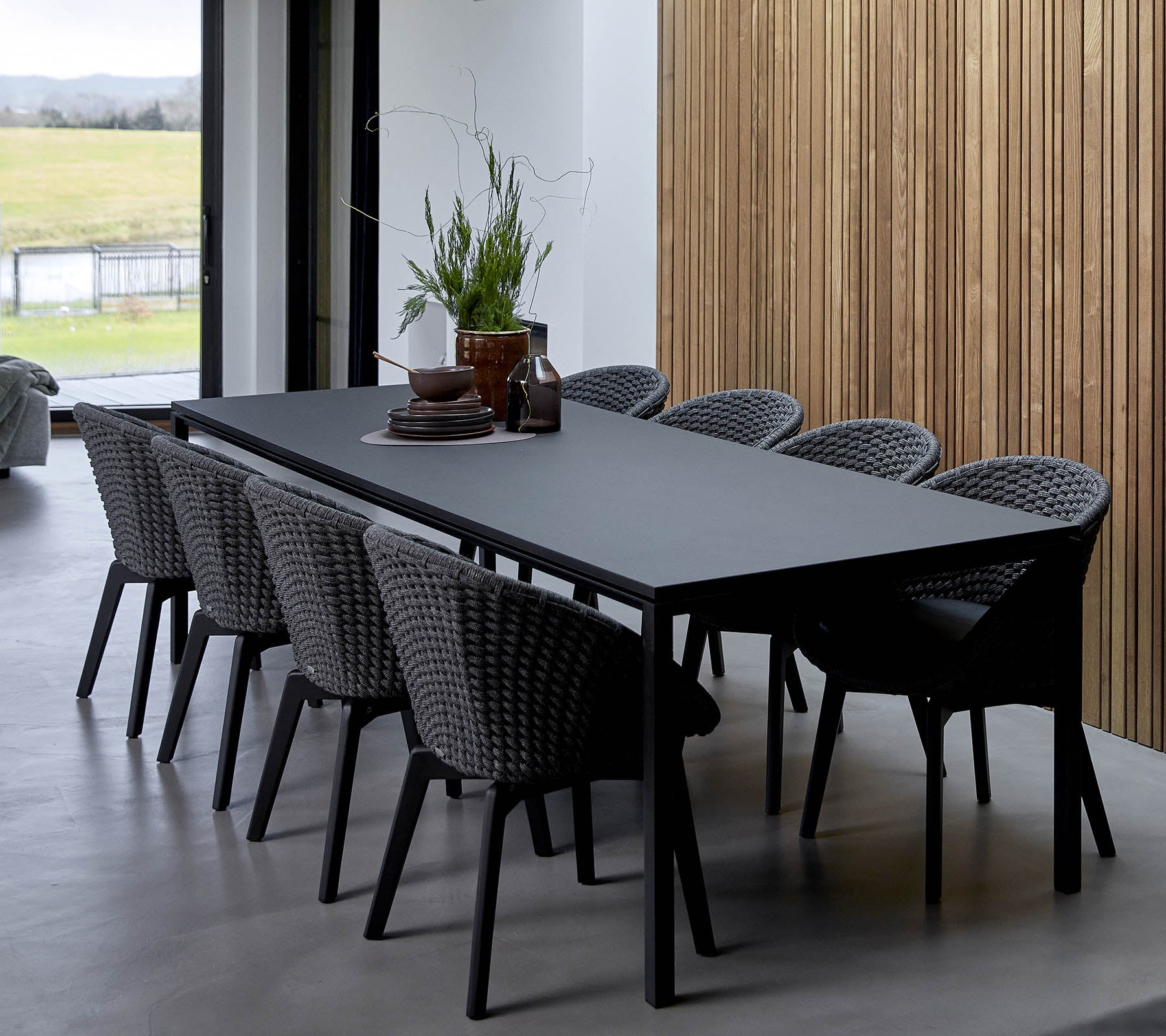 Una mesa elegante rodeada de sillas trenzadas, adornada con plantas decorativas y un centro de mesa, contra un fondo de madera.