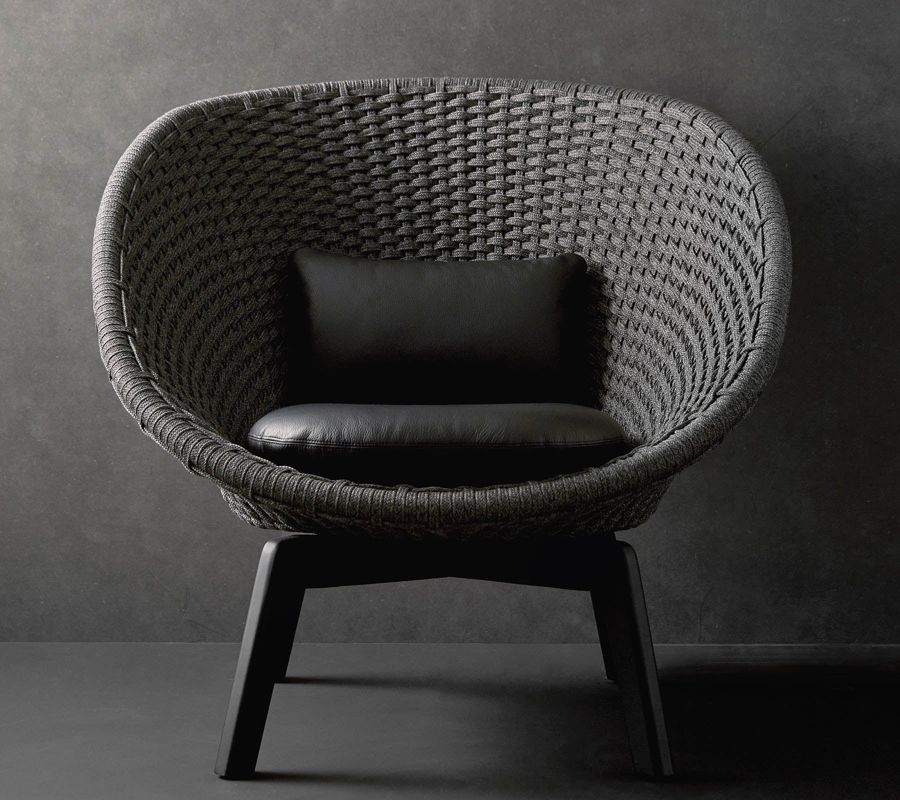 Una silla redonda elegante con un diseño tejido texturizado y un cojín negro, descansando sobre patas de madera elegantes contra un fondo oscuro.
