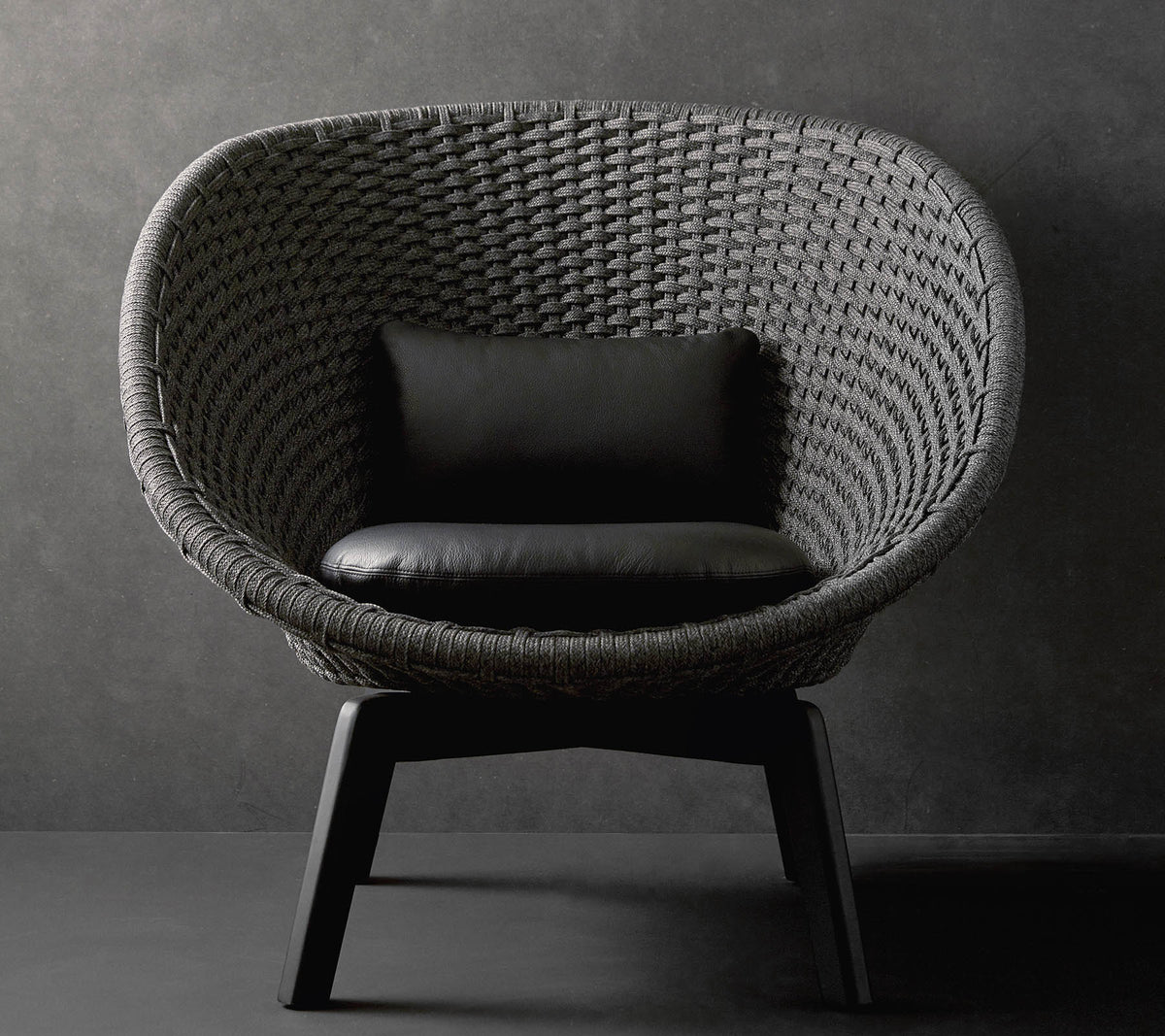 Una silla redonda elegante con un diseño tejido texturizado y un cojín negro, descansando sobre patas de madera elegantes contra un fondo oscuro.