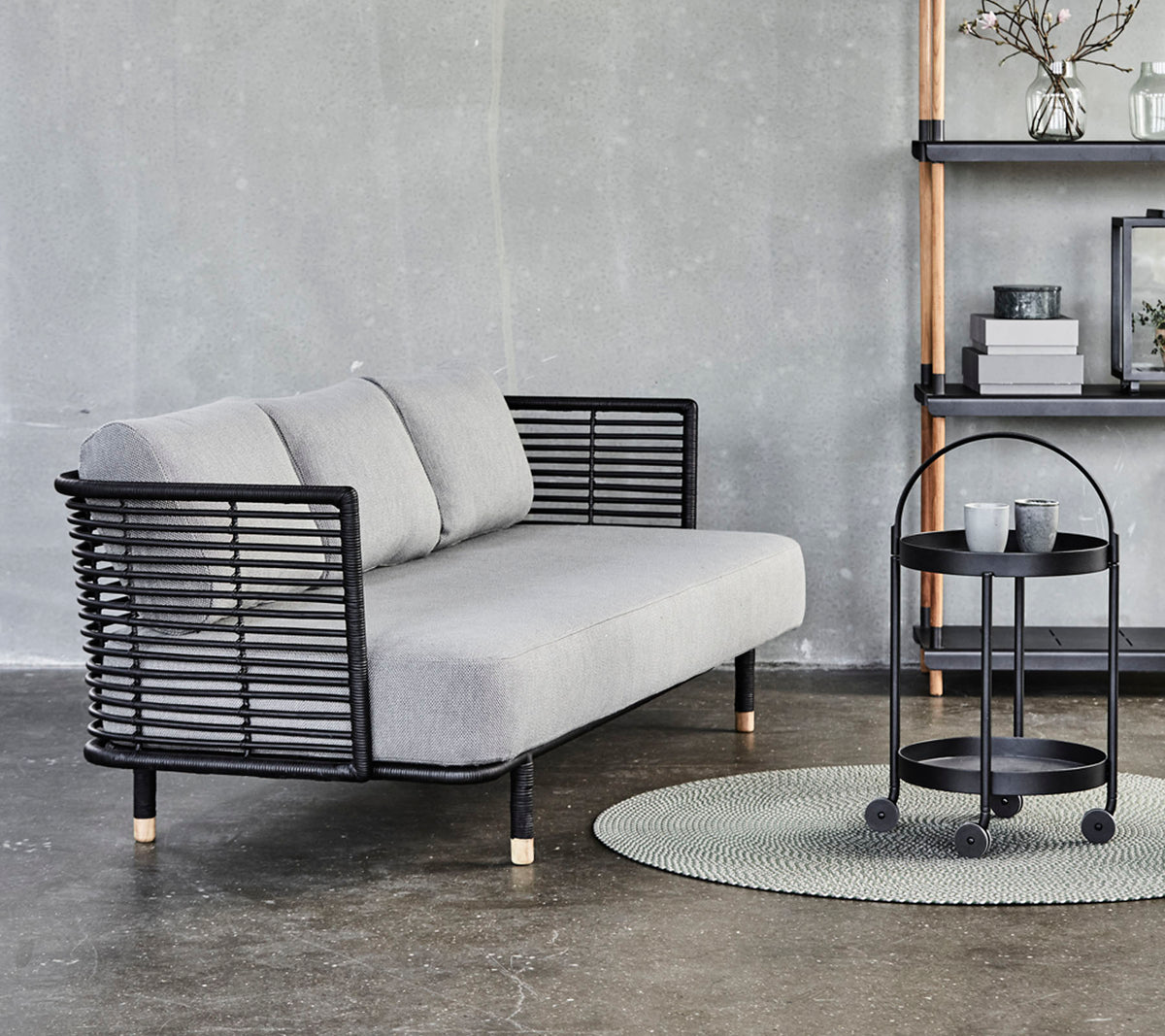 Sofá gris elegante con acentos de metal negro y cojines, acompañado de una mesa lateral redonda sobre una alfombra texturizada contra un fondo neutro.