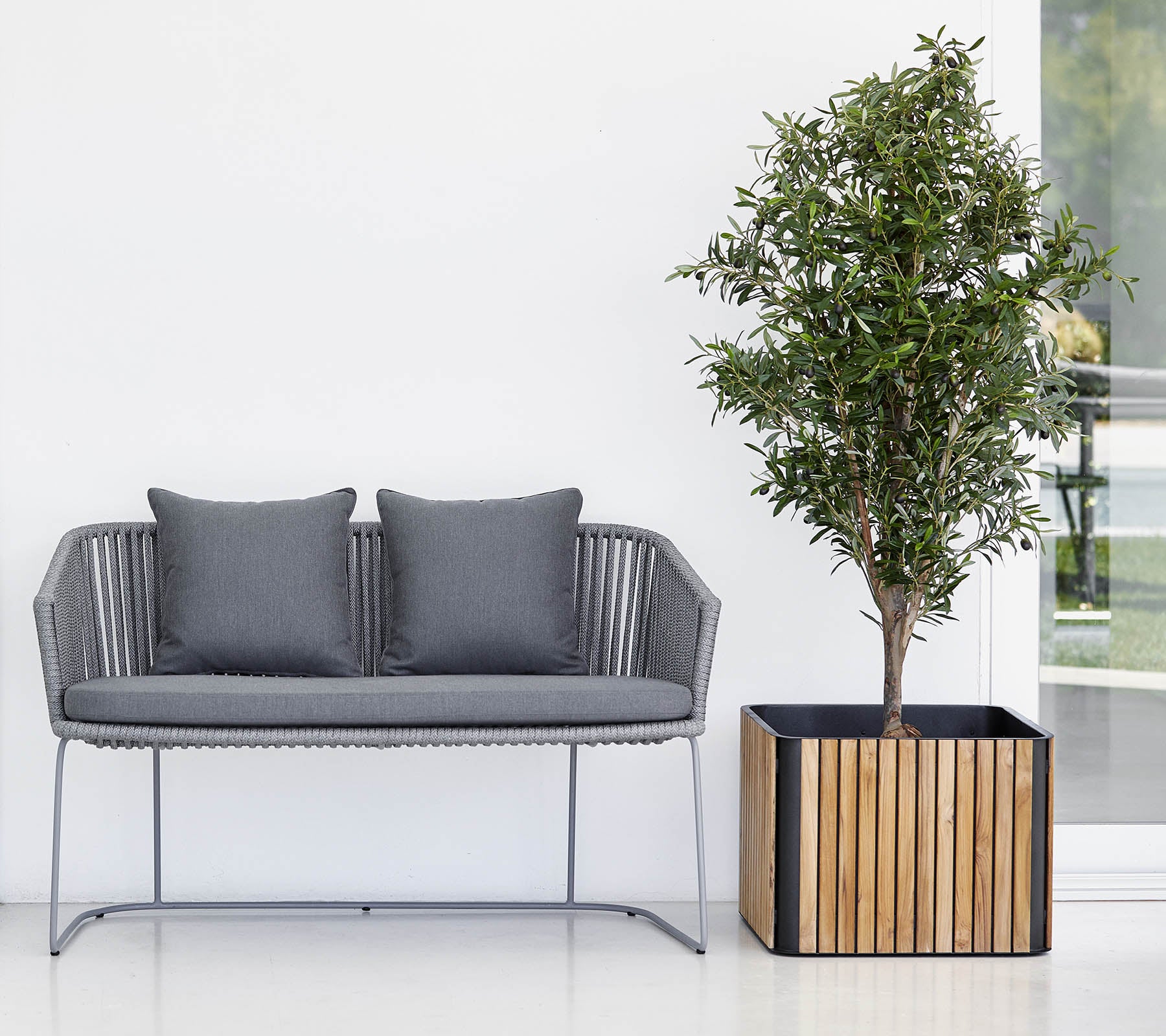 Sofá moderno con dos cojines al lado de una planta en maceta en una maceta decorativa, contra un fondo limpio y neutral.