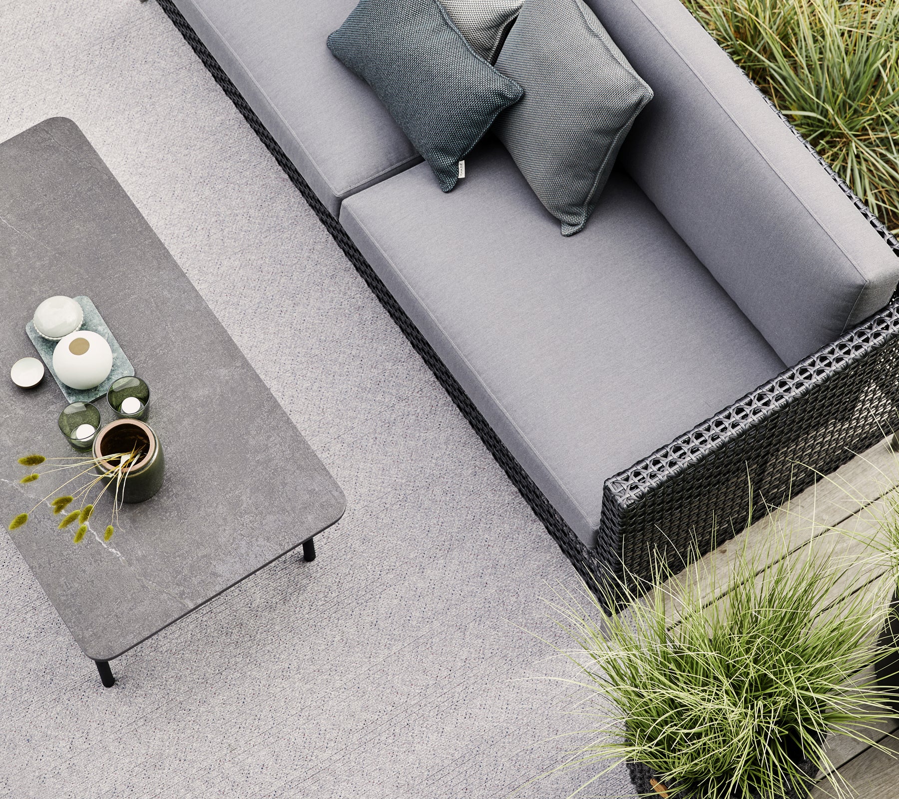 Un sofá gris con dos cojines se encuentra cerca de una mesa minimalista adornada con cerámica y una pequeña planta, rodeada de vegetación.