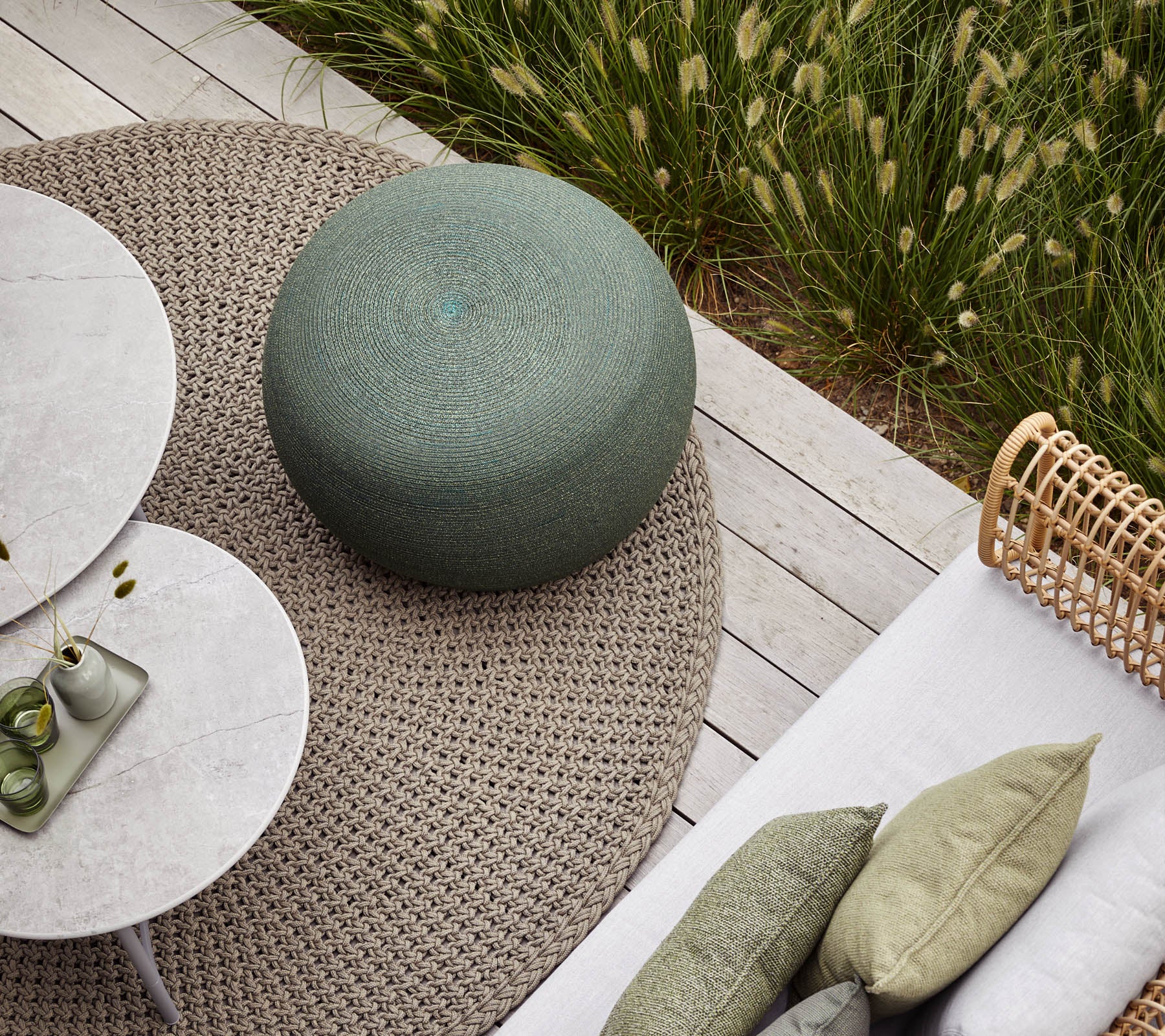 Una acogedora área de asientos al aire libre con un pouf verde redondo, mesas circulares y cojines texturizados sobre una alfombra neutral. Vegetación exuberante al fondo.