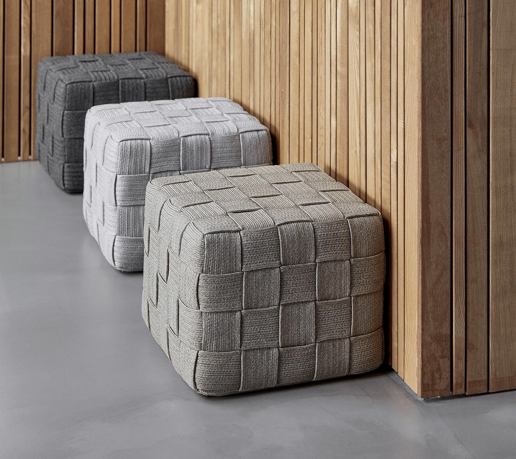 Tres ottomanes texturizados y cuadrados en tonos de gris variados están posicionados contra una pared de madera, añadiendo un toque moderno al entorno.