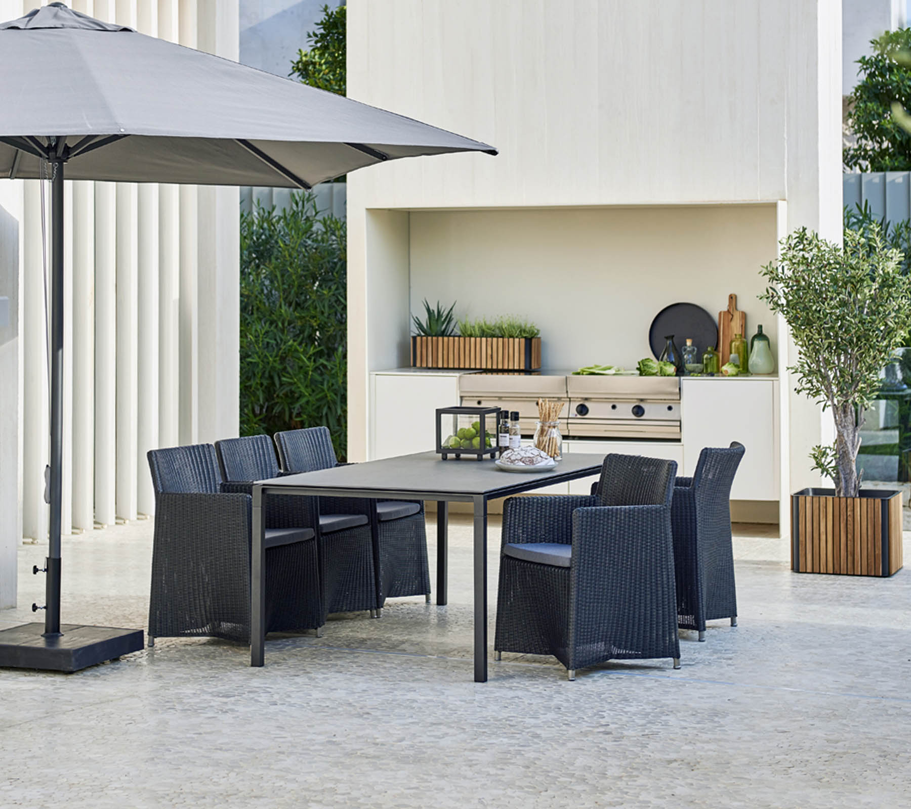 Conjunto de comedor exterior moderno con una gran mesa, seis sillas, una sombrilla elegante y un fondo con vegetación y elementos decorativos.
