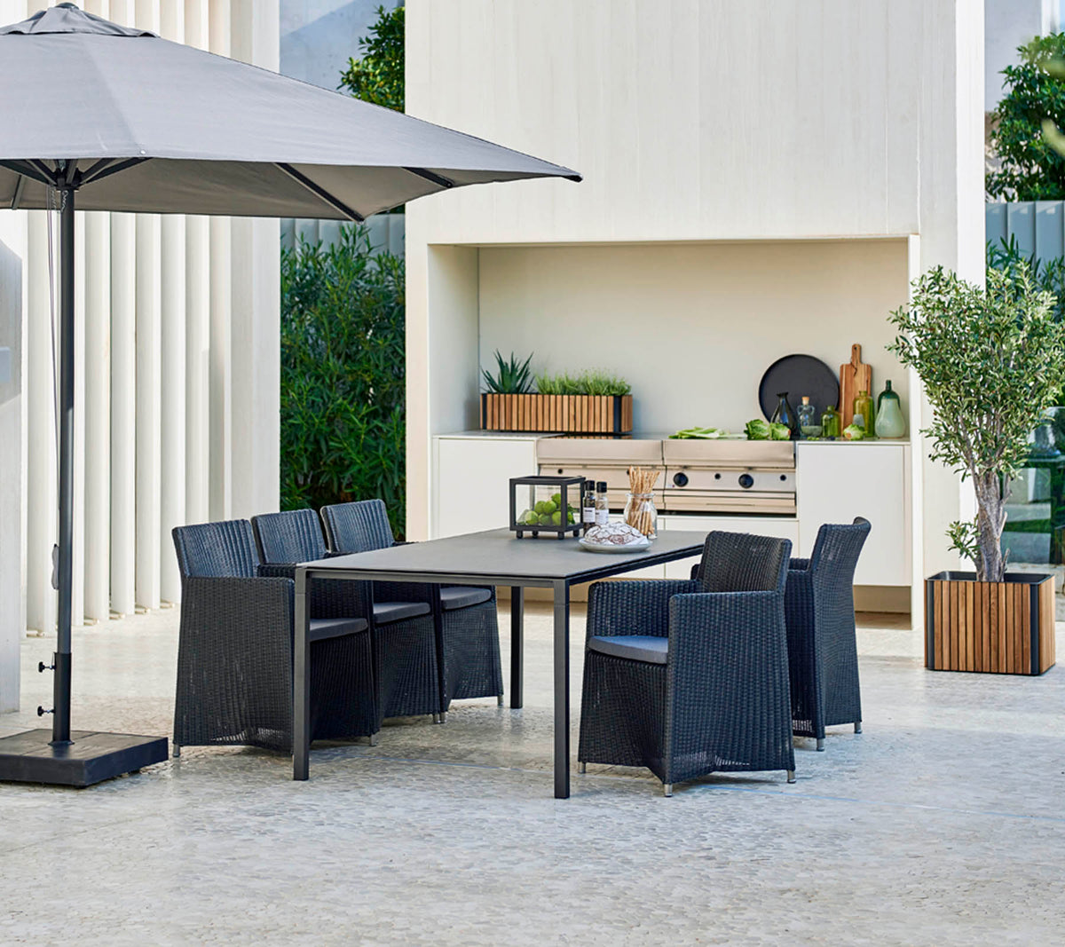 Conjunto de comedor exterior moderno con una gran mesa, seis sillas, una sombrilla elegante y un fondo con vegetación y elementos decorativos.
