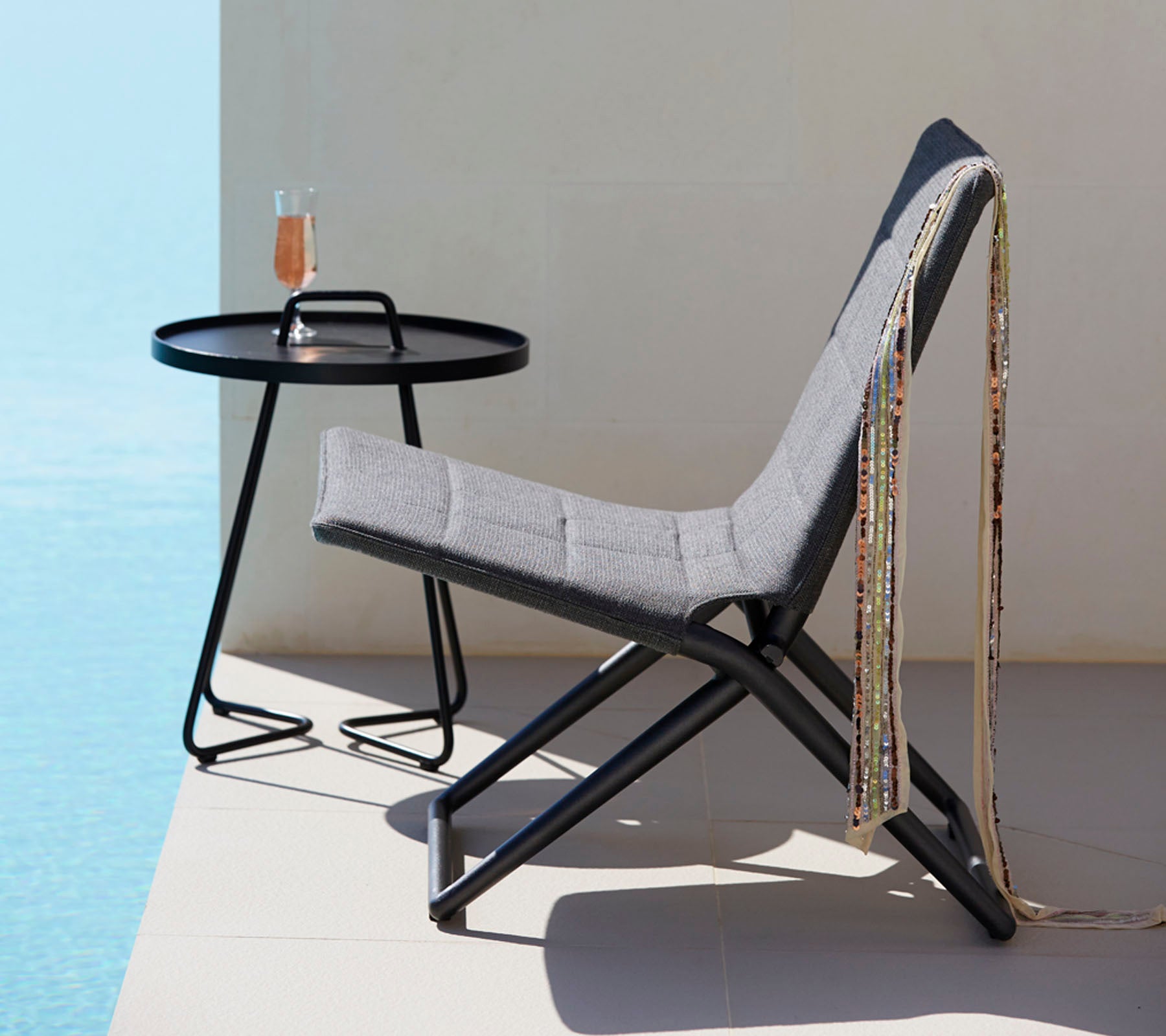 Una silla elegante con tela gris y un marco de metal negro elegante, acompañada de una mesa redonda que sostiene una bebida y cuentas decorativas.