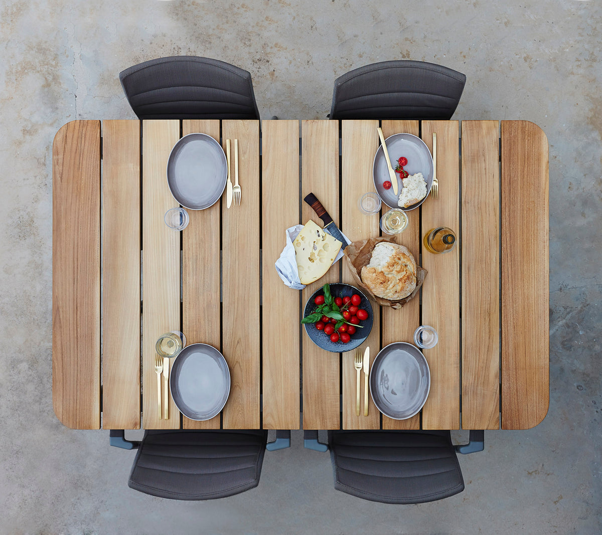 Una mesa de madera servida con platos, vasos y una variedad de quesos, frutas y pan, rodeada de cuatro sillas.