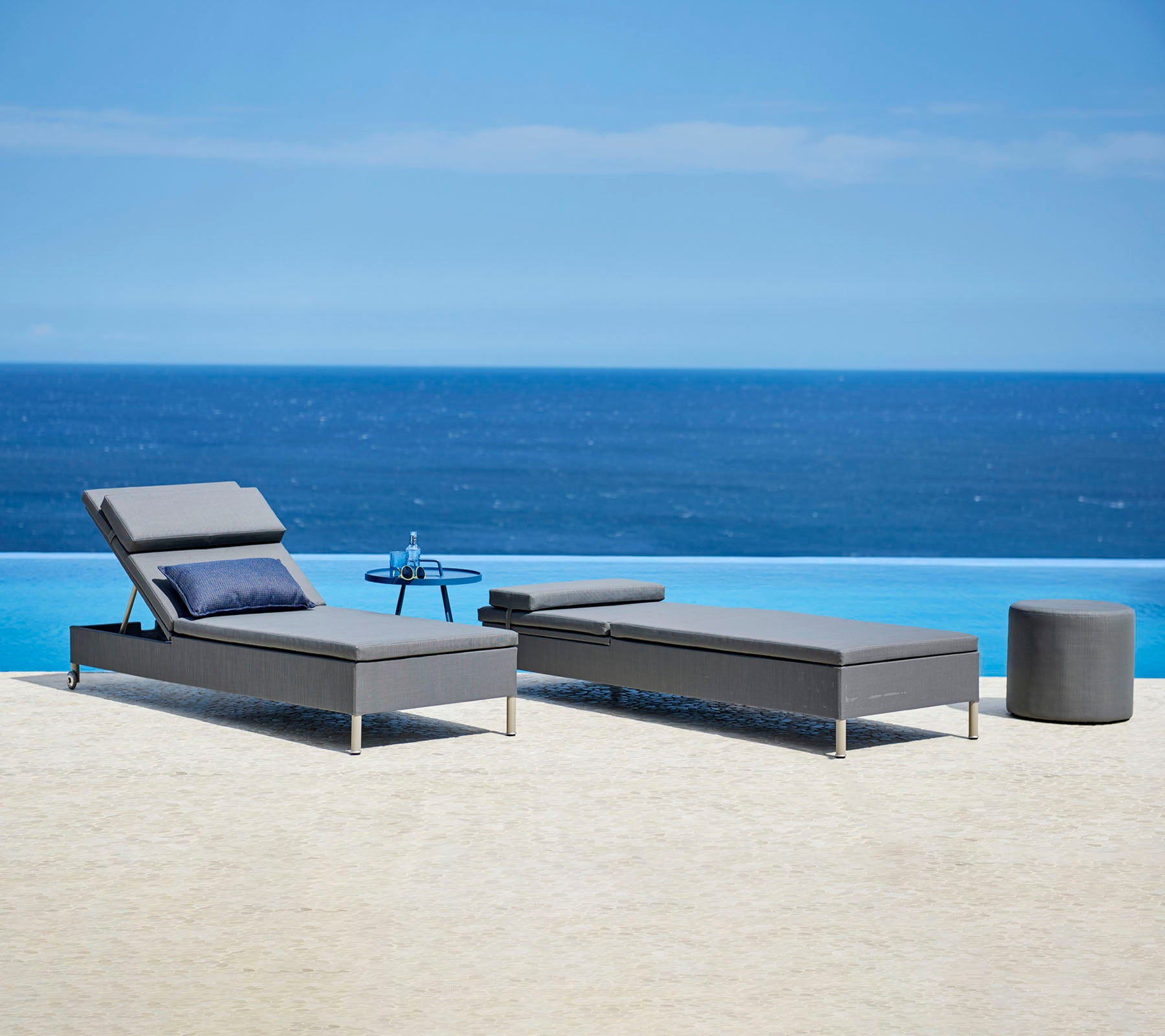 Dos elegantes tumbonas grises con cojines y una pequeña mesa auxiliar junto a una tranquila fuente de agua azul, perfectas para relajarse.