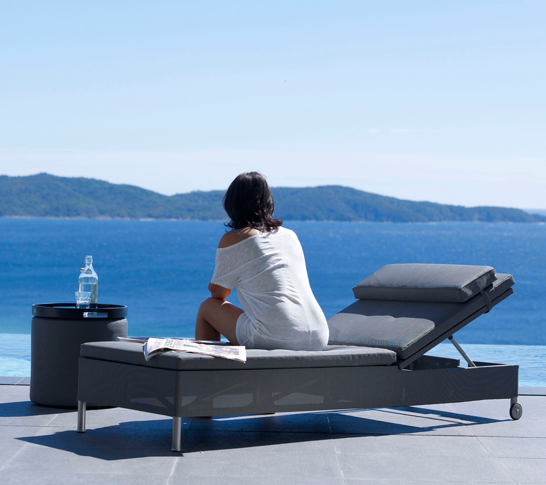 Una mujer sentada en una silla de lounge, mirando una serena vista del océano, con una botella de agua en una pequeña mesa a su lado.