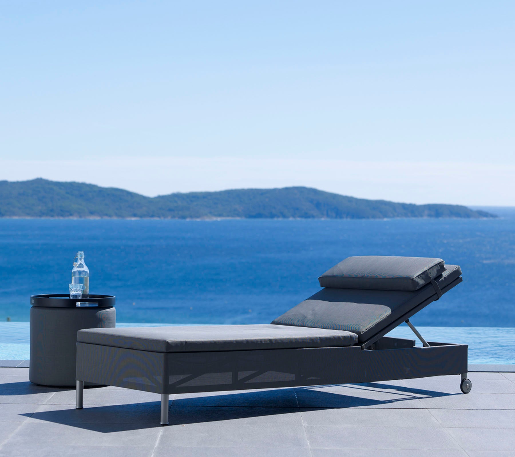 Una silla de lounge moderna y una mesa lateral junto a un sereno cuerpo de agua con colinas distantes bajo un cielo despejado, perfecto para relajarse.