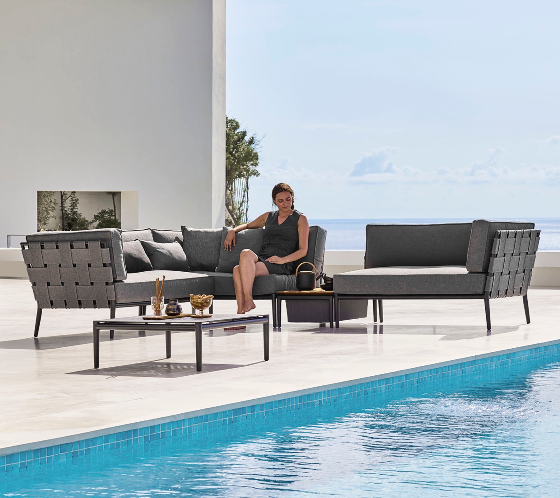 Un elegante conjunto de salón al aire libre con asientos elegantes y una mesa de café de vidrio junto a una piscina, con una persona relajándose cómodamente.
