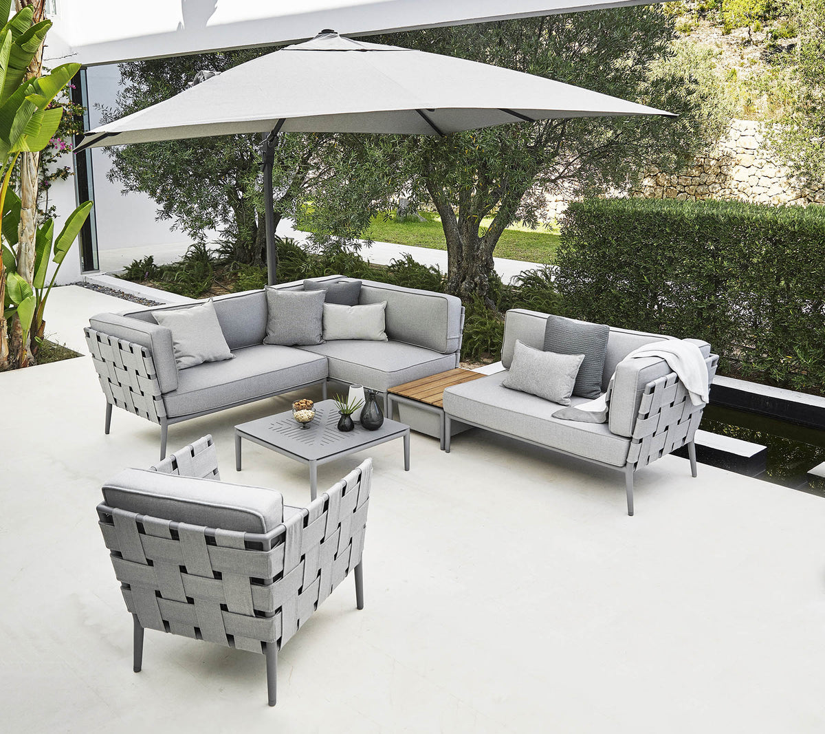 Arreglo de asientos al aire libre elegante con un sofá seccional gris, una silla y una mesa de café bajo una gran sombrilla.