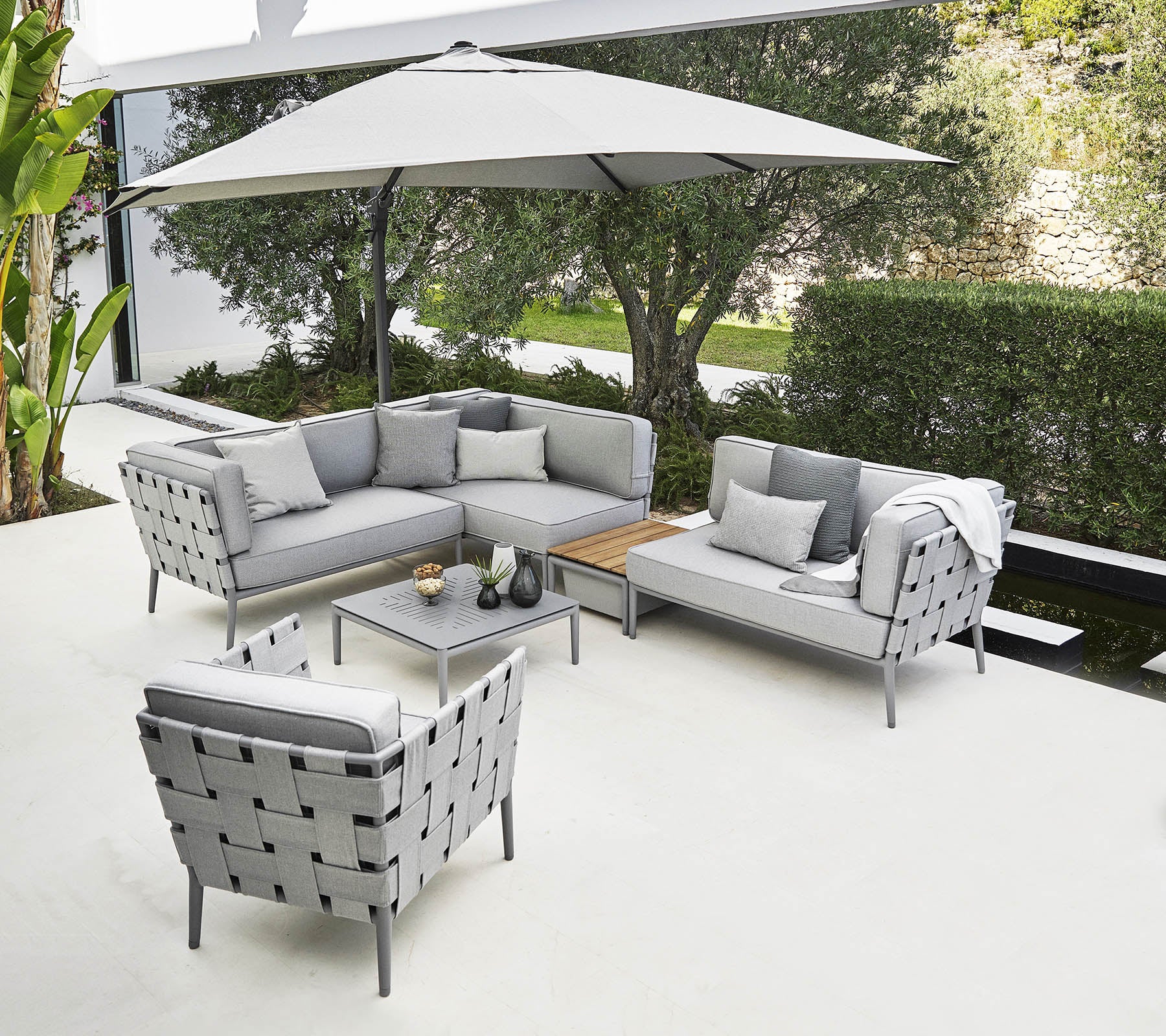 Arreglo de asientos al aire libre elegante con un sofá seccional gris, una silla y una mesa de café bajo una gran sombrilla.